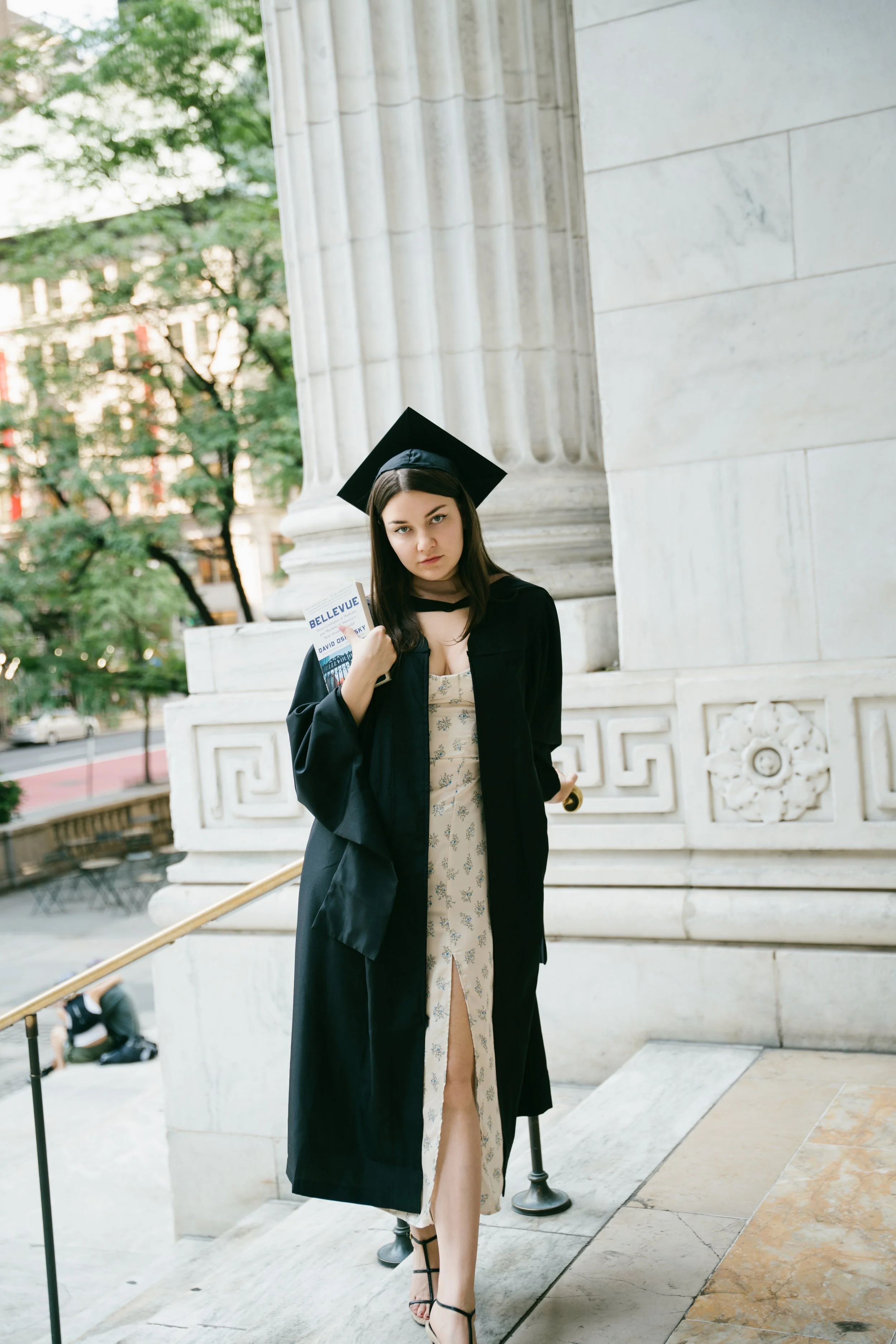 Young woman in graduation gown and cap standing outside near a stone building, holding a book titled 'BELLVUE', with trees and buildings in the background.