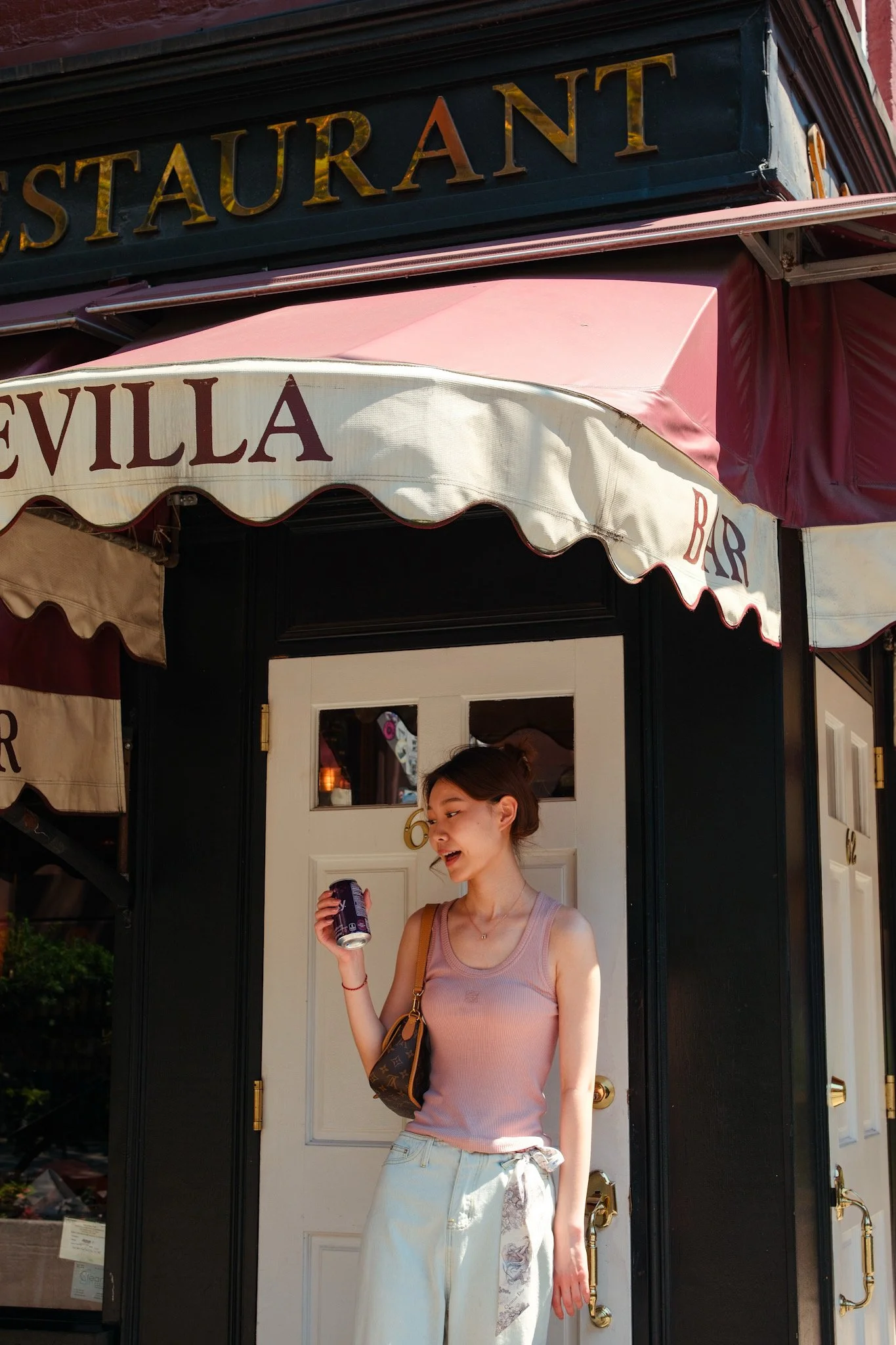 Young woman with light skin, wearing a pink tank top and light-colored pants, holding a soda can and smiling, standing in front of a restaurant storefront with black and white doors, and signage indicating a bar and restaurant.