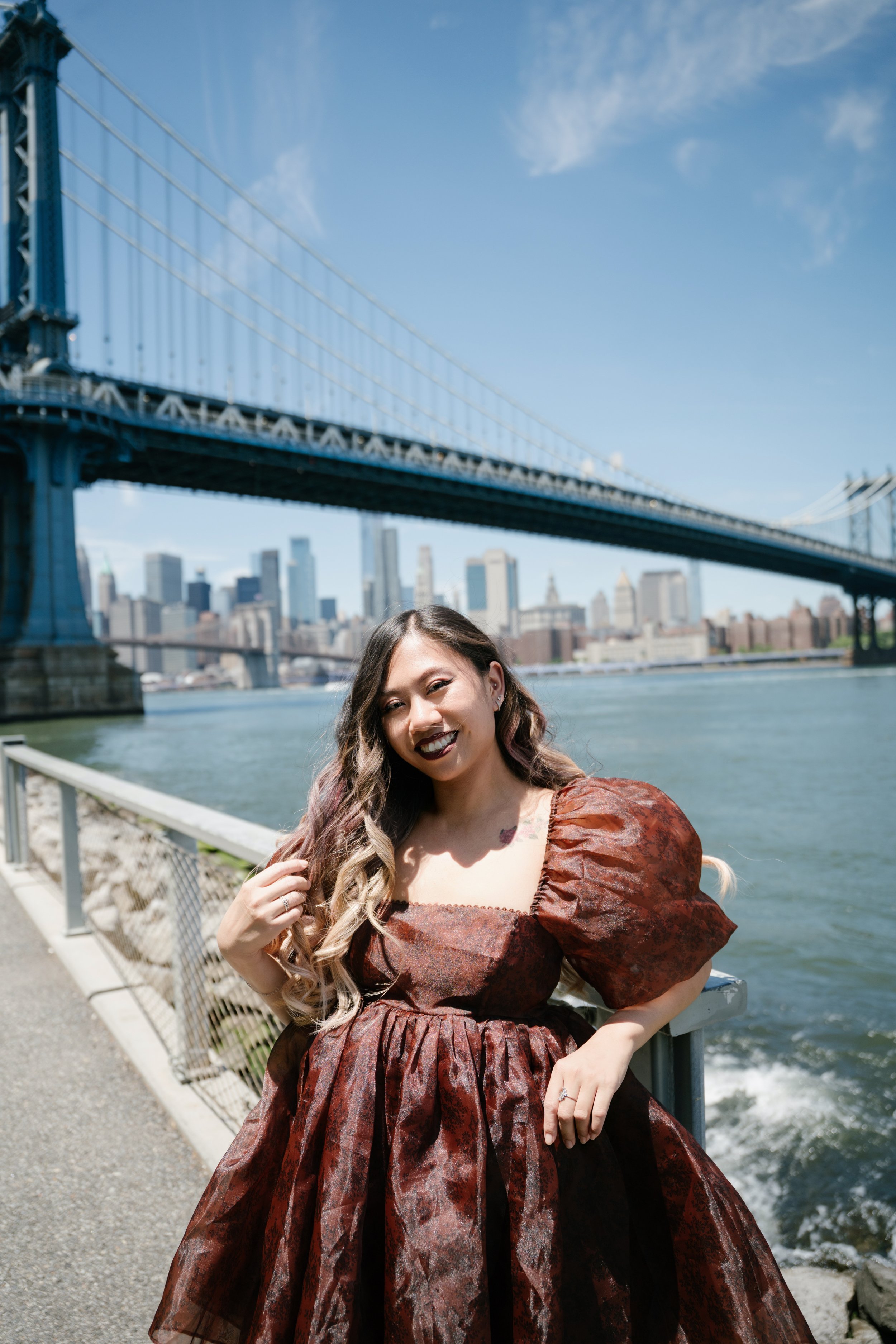 A woman with long wavy hair smiling, wearing a patterned brown and black dress with puffed sleeves, standing near the water with the Brooklyn Bridge and Manhattan skyline in the background.