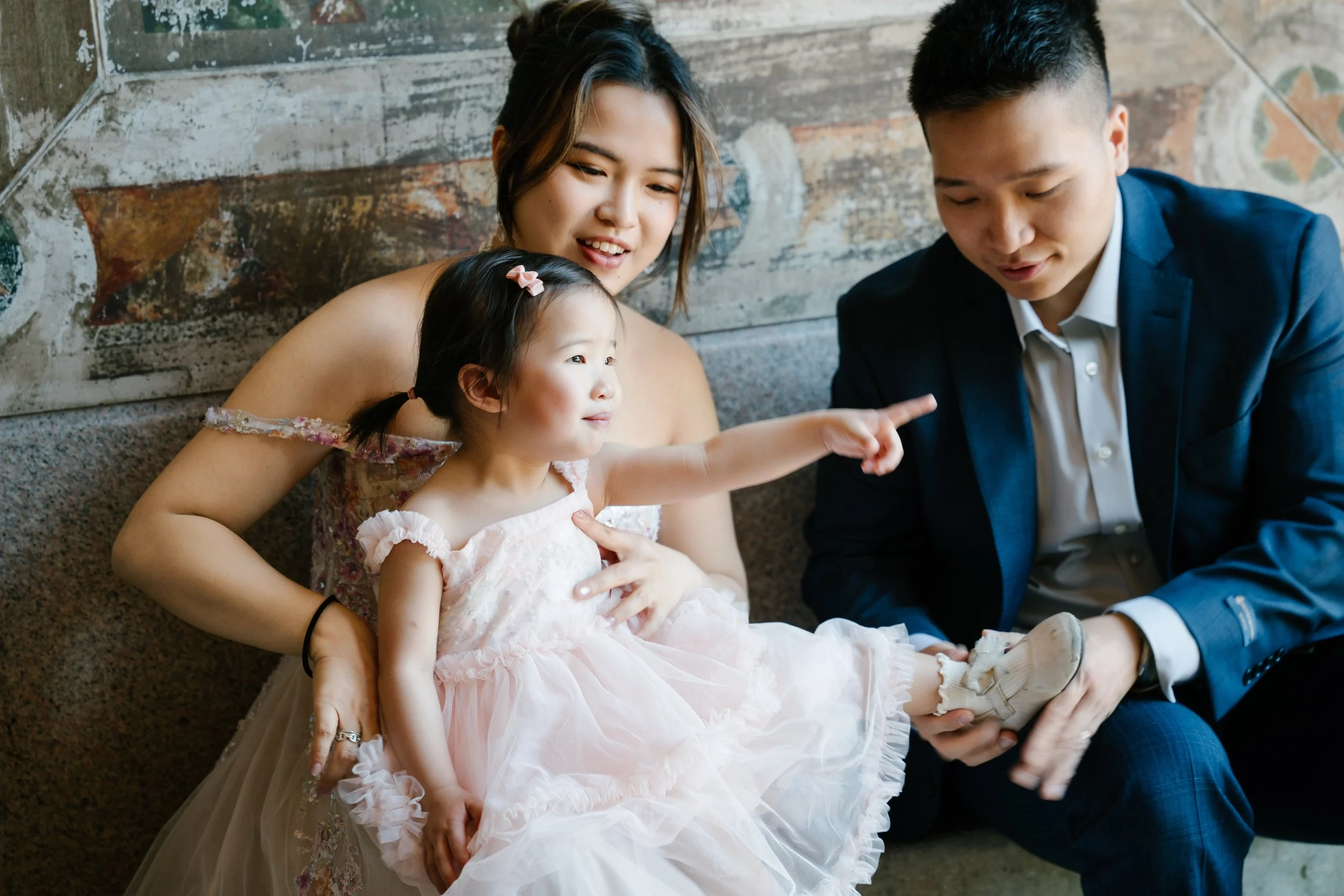 A young girl in a pink dress sits on a man's lap, pointing at him with her finger. A woman with blonde hair looks at the girl with a smile. The man, dressed in a blue suit, is looking at the girl. They are sitting on the floor with a rustic, wooden w