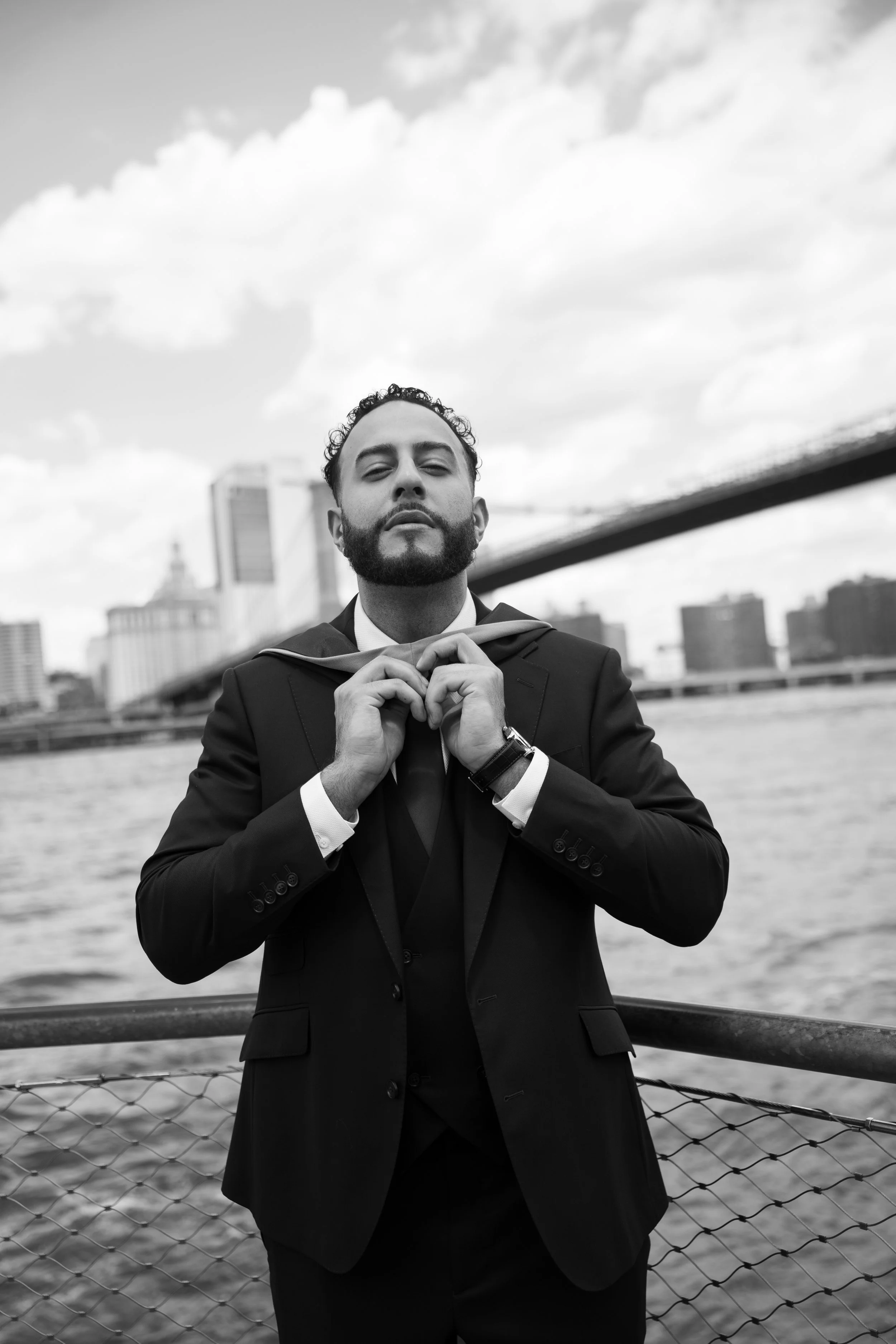 A man in a suit standing by a waterfront, adjusting his tie, with a city skyline and bridge in the background.