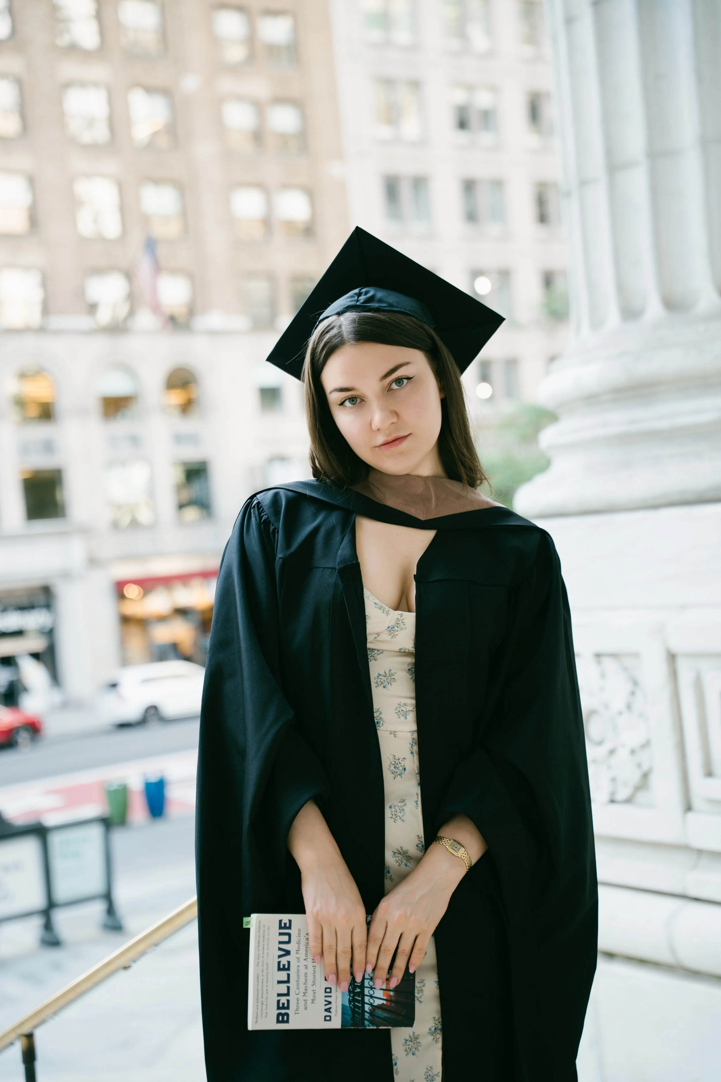 A young woman in a graduation cap and gown stands on a porch holding a book titled 'Belleville.' She is looking at the camera with a serious expression. There is a cityscape with tall buildings in the background.