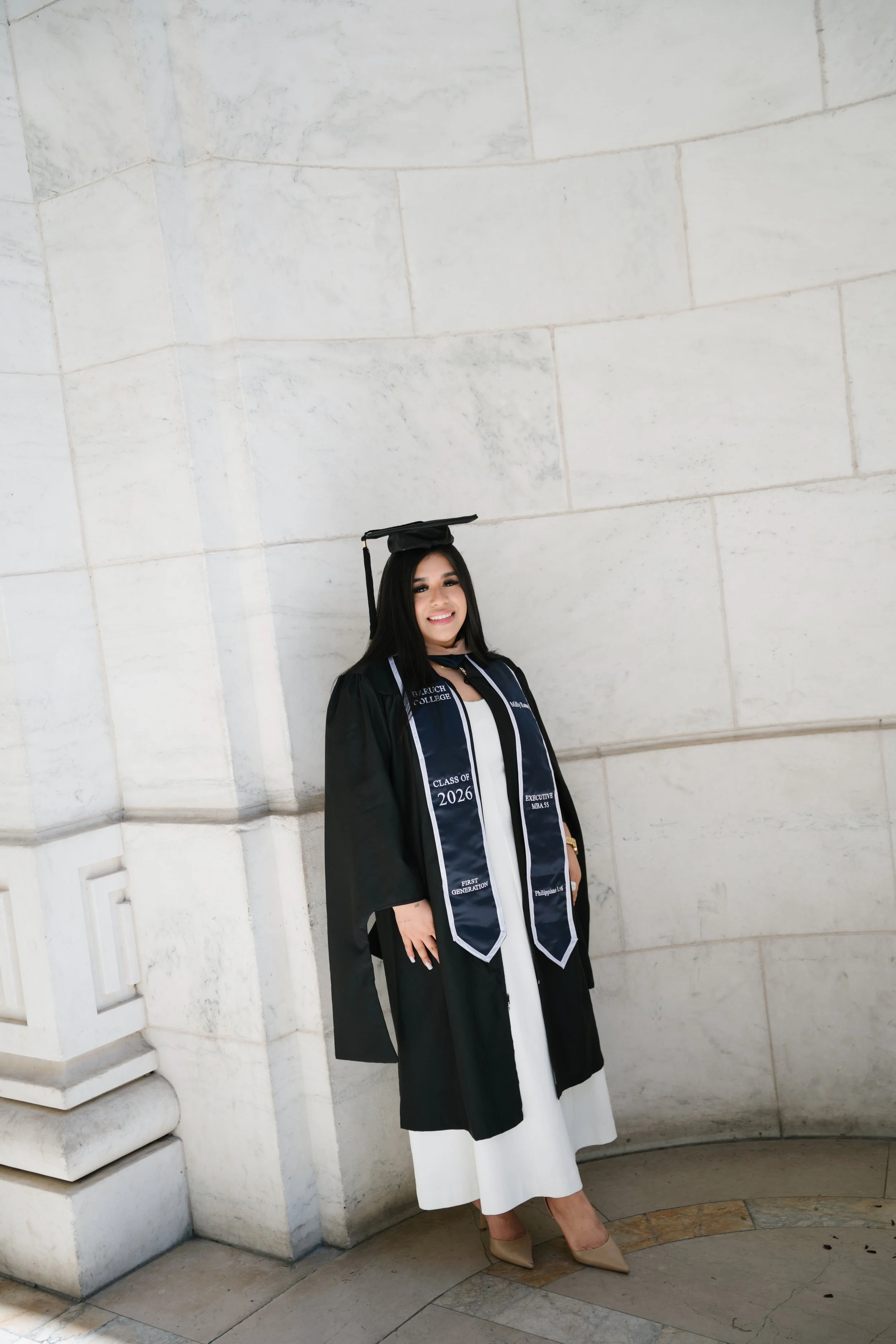 A woman in graduation attire, including a black cap and gown with a sash, standing against a white marble wall, smiling at the camera.