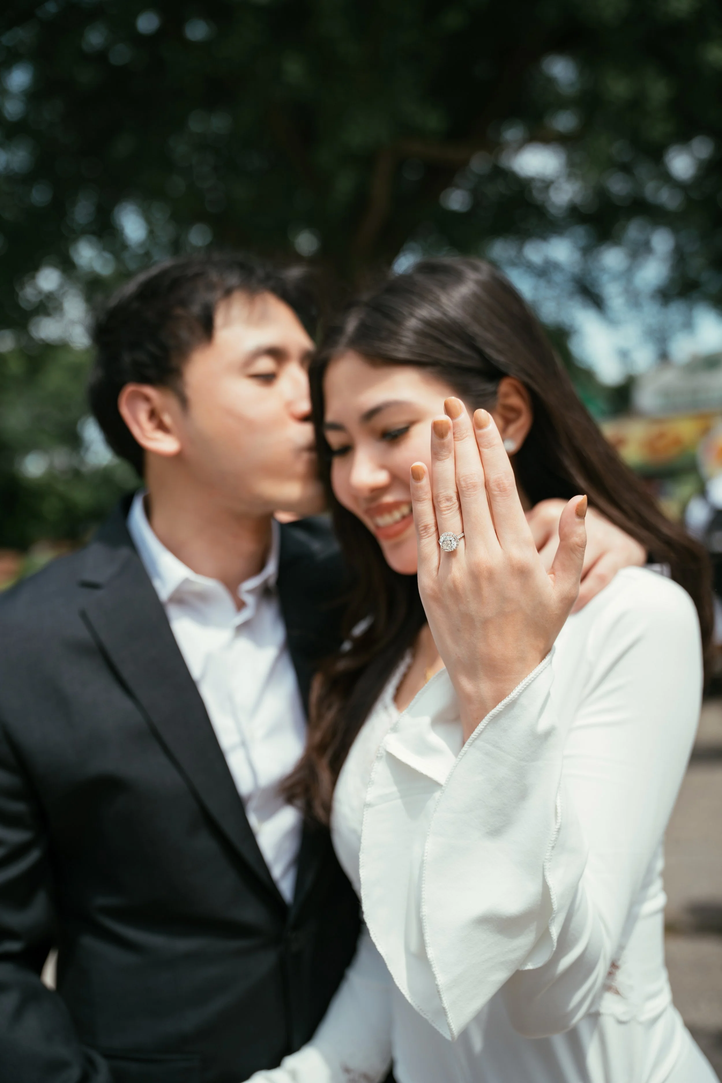 A couple dressed in formal attire, with the man whispering into the woman's ear and the woman showing off her engagement ring with a big diamond, smiling.