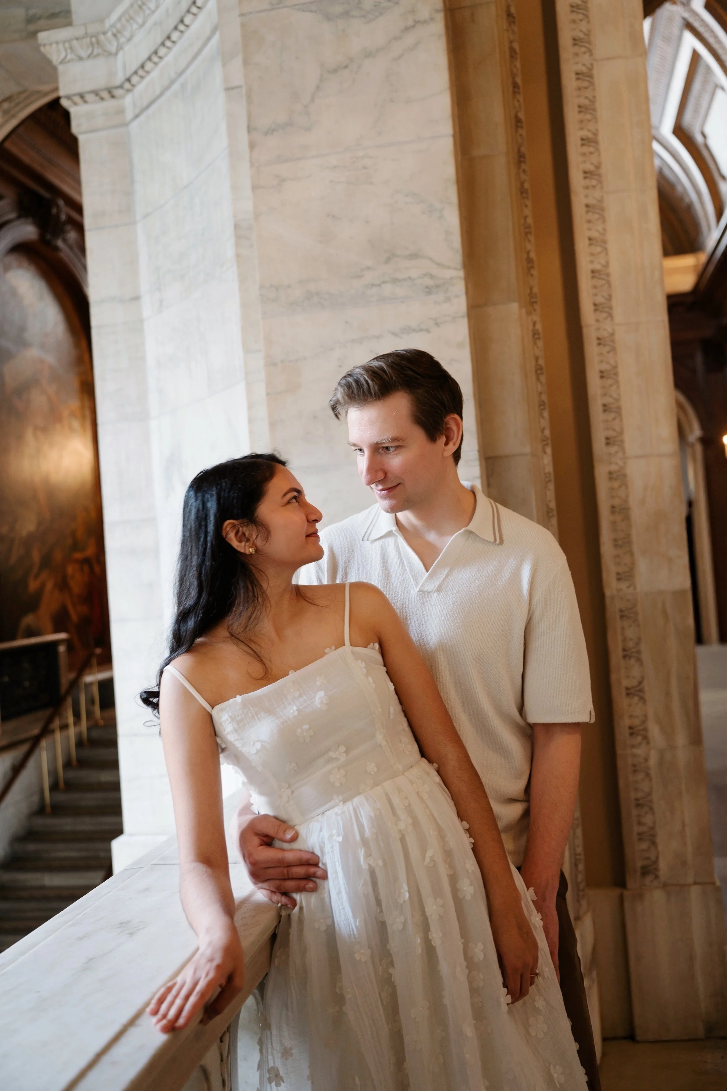 A young couple stands close together in a historic building with ornate architecture. The woman has long dark hair and is wearing a white dress with floral embroidery. The man is taller, with short brown hair, and is wearing a light-colored shirt. Th