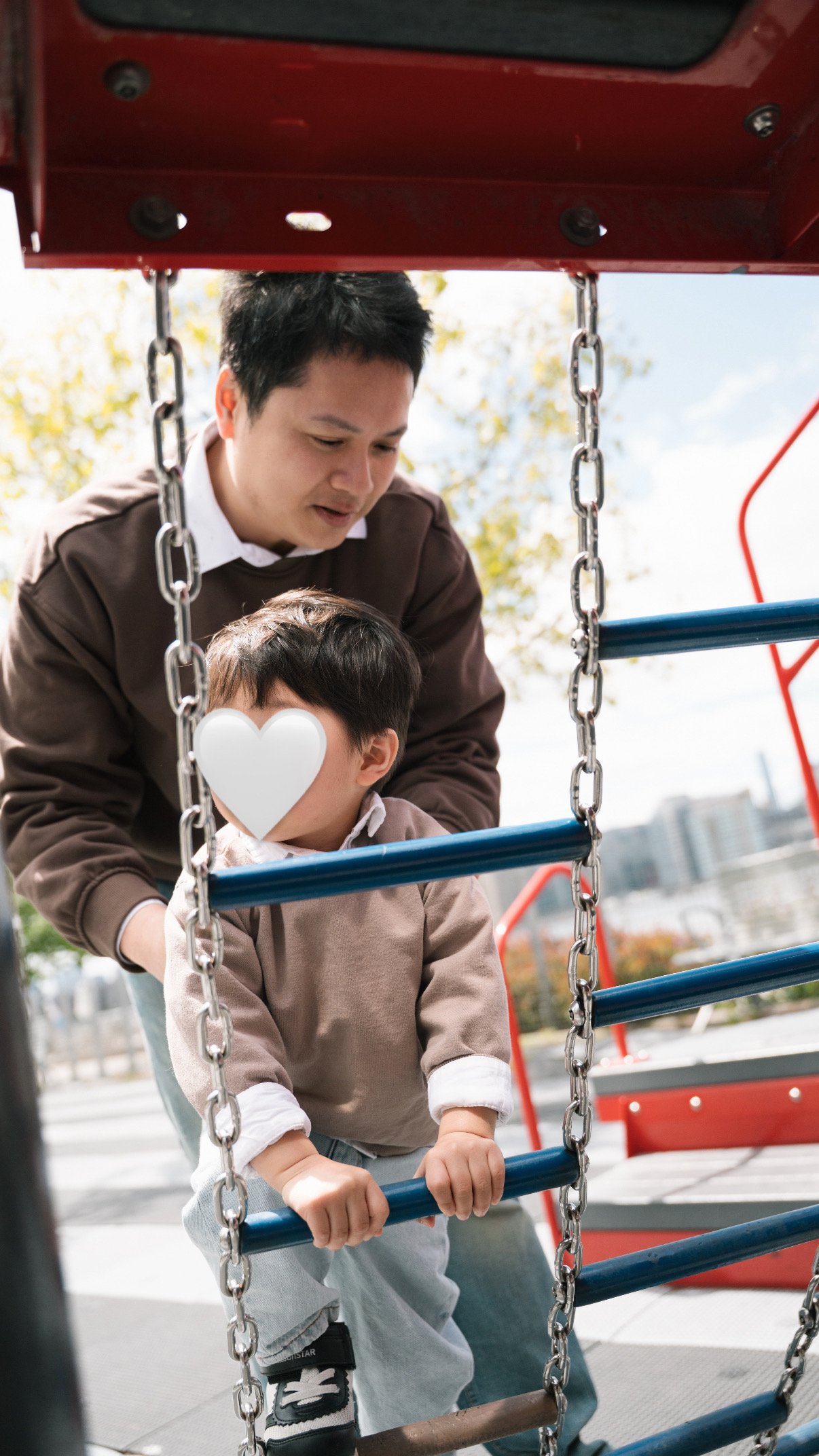 A man helping a young boy on a playground climbing structure with blue bars and metal chains, outdoors on a sunny day with trees and a building in the background.