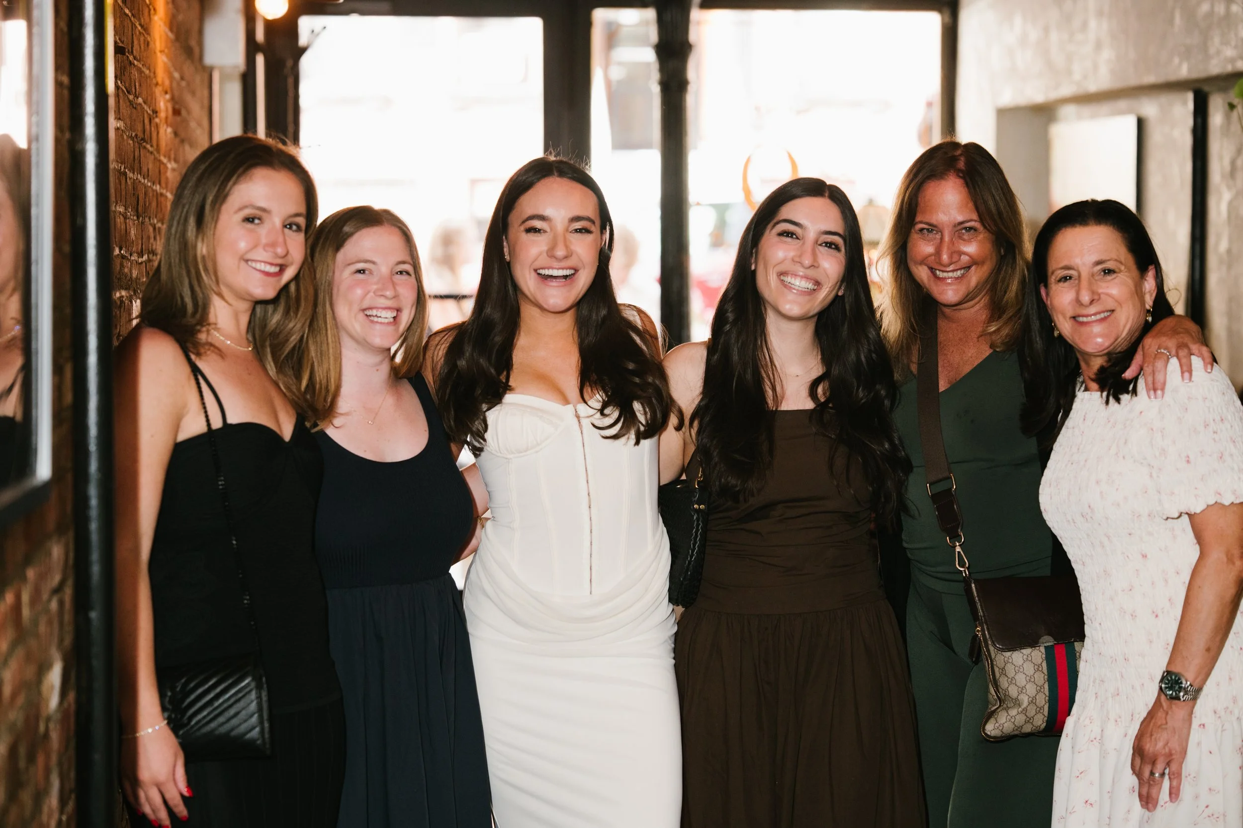 A group of seven women standing together indoors, smiling for the camera. They are dressed stylishly and appear to be at a social gathering.