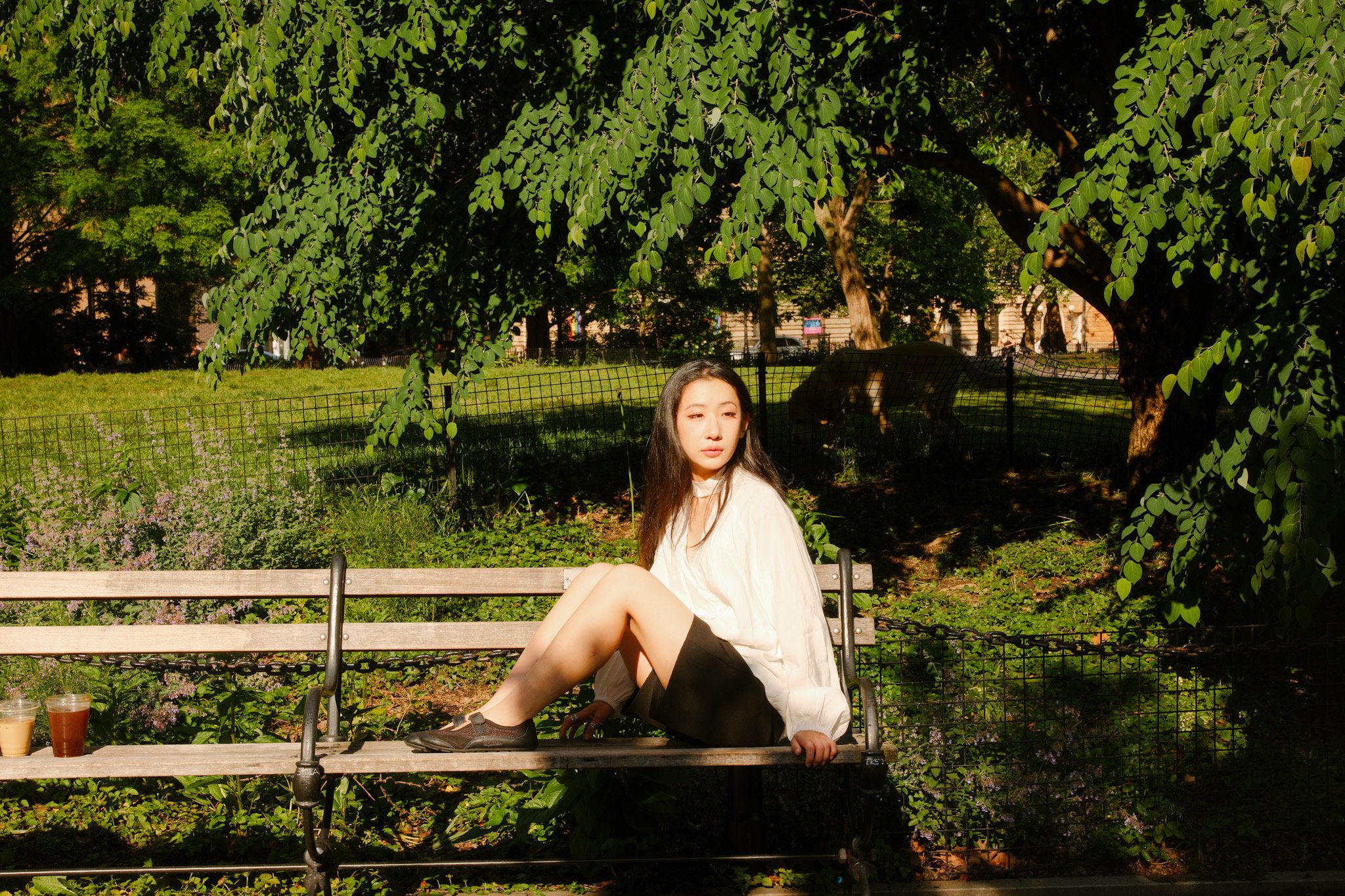 A young woman with long dark hair sitting on a park bench in sunlight, surrounded by greenery and trees, with two drinks placed on the bench beside her.
