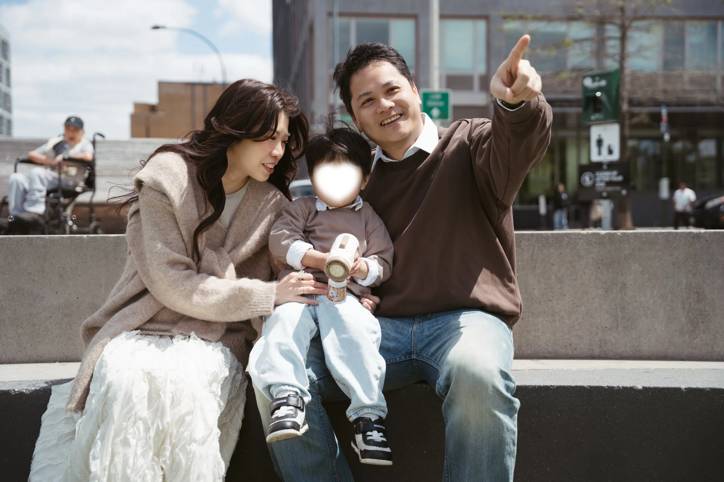A smiling family of three sitting on a bench outdoors in an urban setting, with an elderly person in a wheelchair in the background.