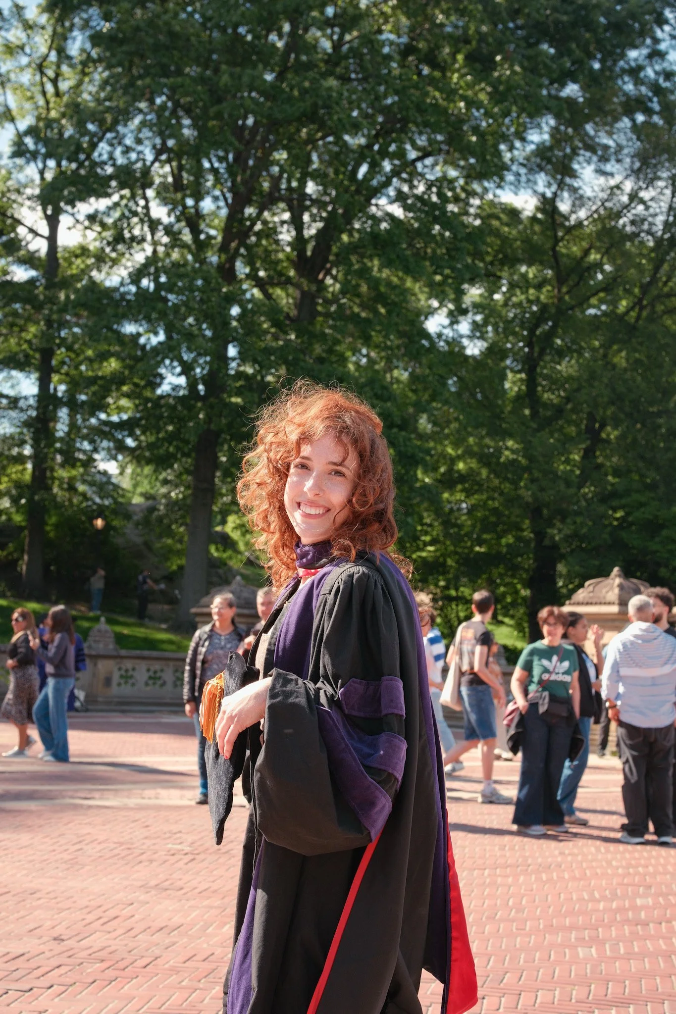 A young woman with curly red hair smiling outdoors, wearing a graduation gown and holding a certificate, with a crowd and trees in the background.