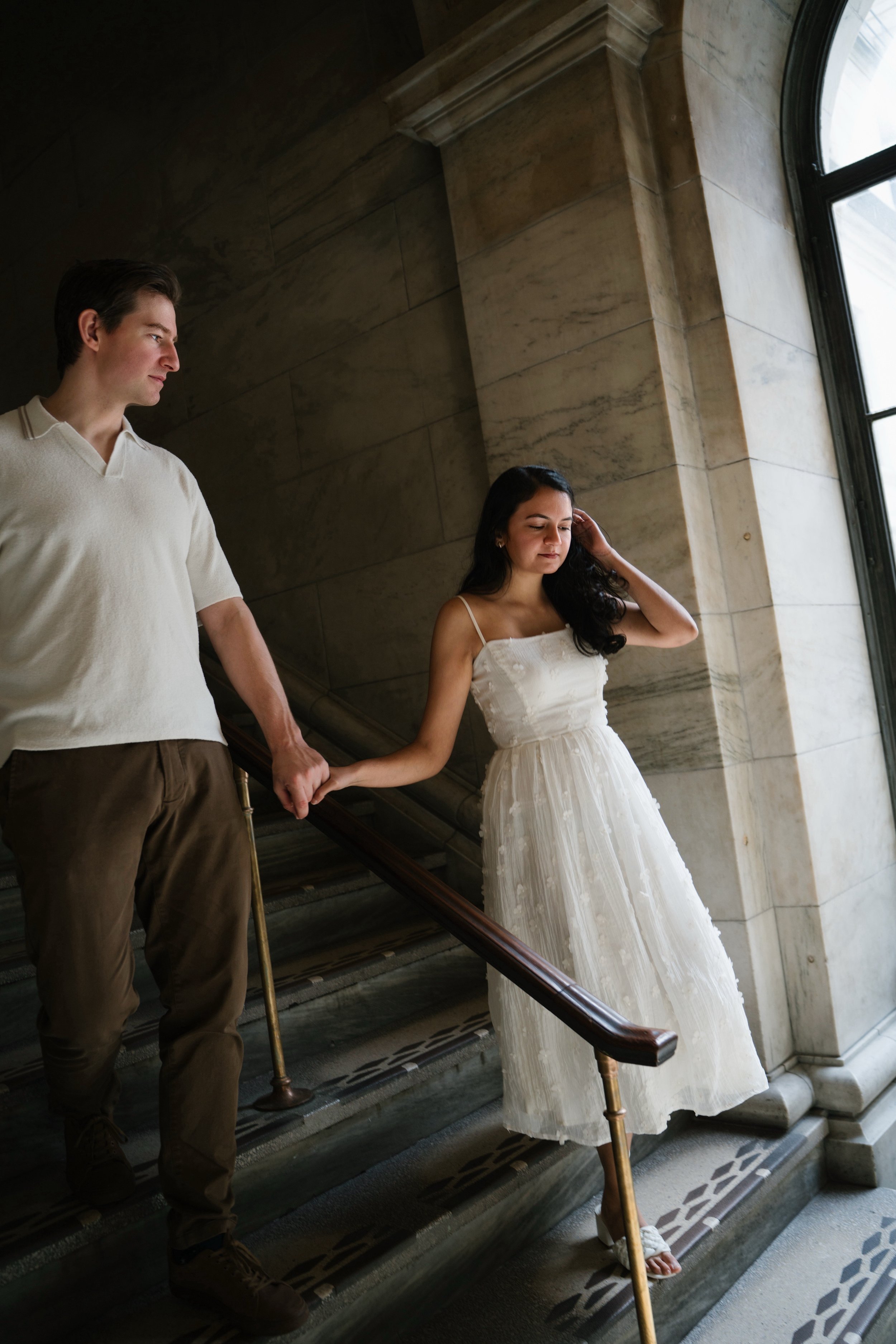 A man and a woman holding hands on a staircase indoors near a large window, with the man wearing a beige shirt and brown pants, and the woman wearing a white dress with a sheer overlay.