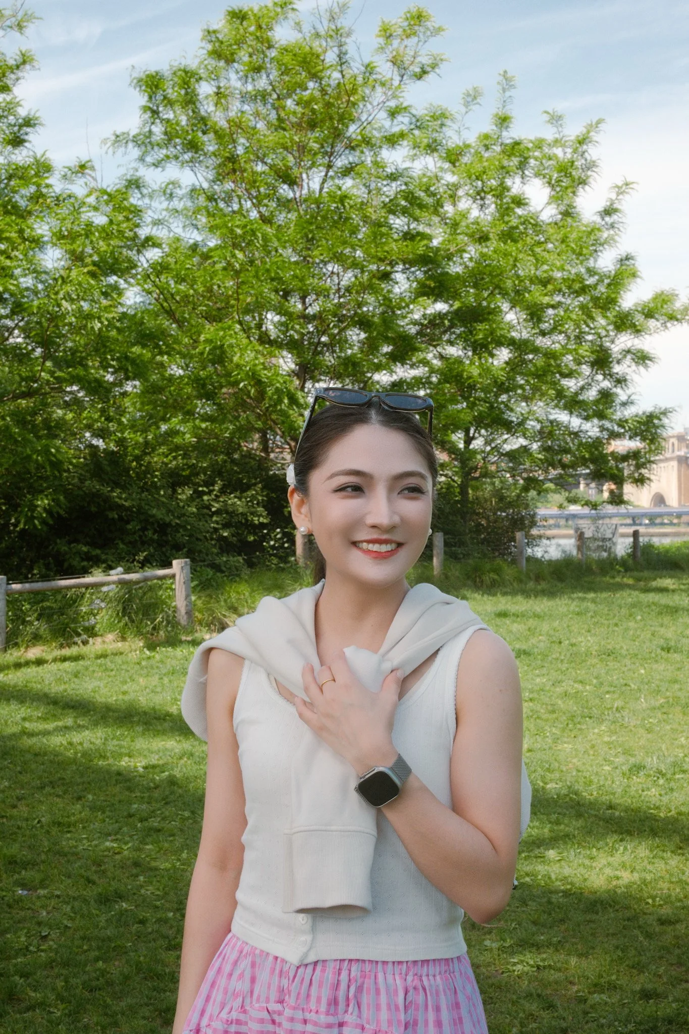 A young woman smiling outdoors in a park, wearing a sleeveless white top and pink striped skirt, with a gray sweatshirt draped over her shoulders, sunglasses on her head, and a smartwatch on her wrist, standing in front of green trees and a fence.