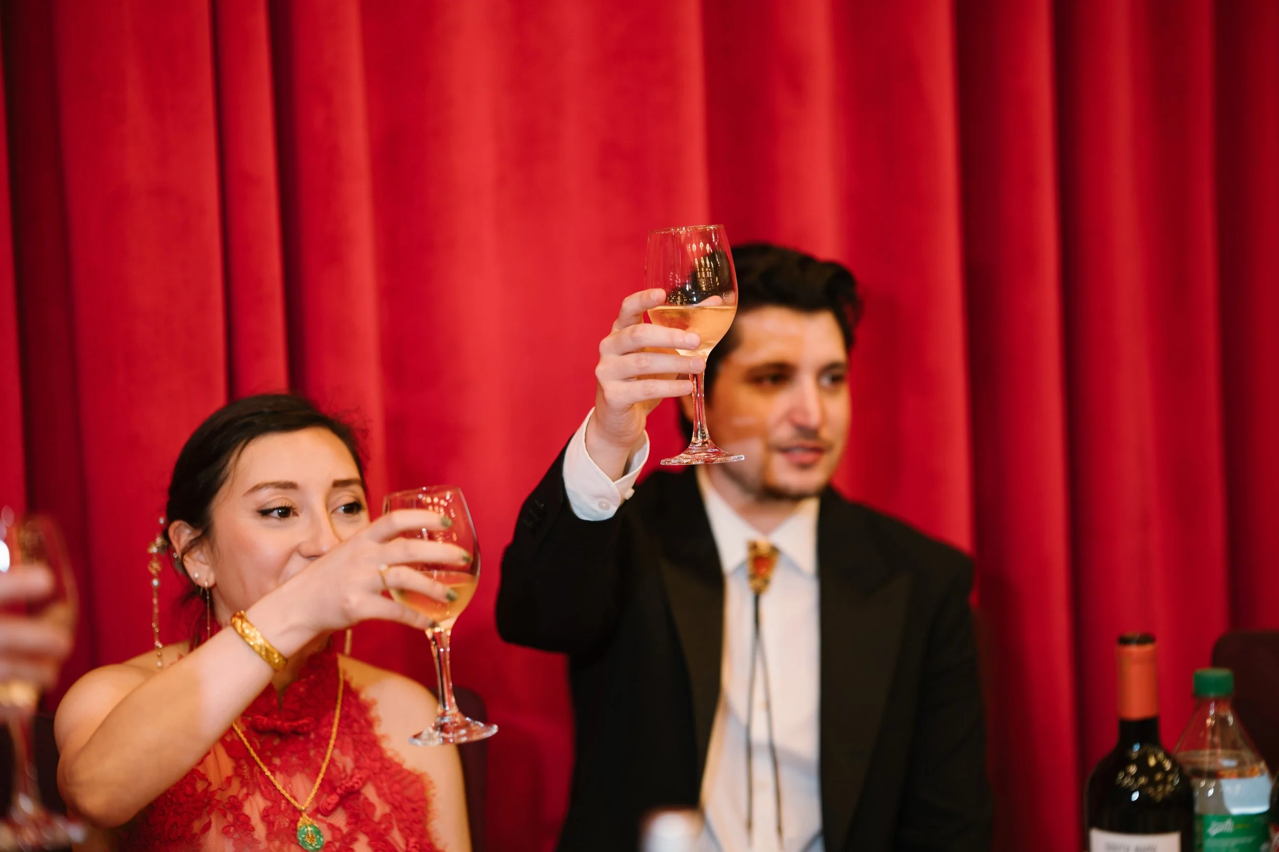 People raising glasses of white wine during a celebration, with a red curtain in the background.