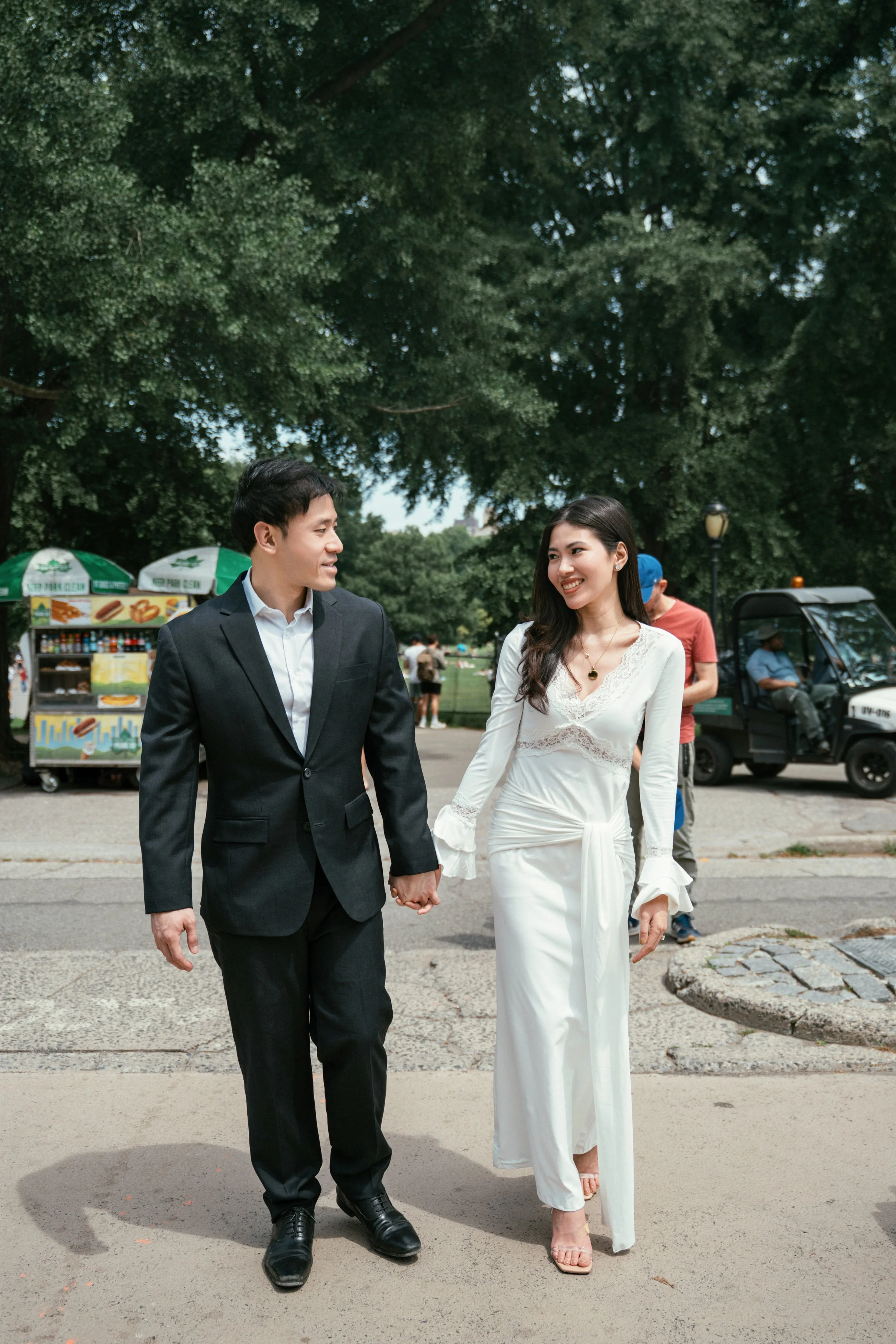 A couple dressed in formal attire holding hands and walking outdoors in a park. The man is wearing a black suit, white shirt, and black shoes, and the woman is in a long white dress with lace details and open-toed sandals. There are trees, a hot dog 