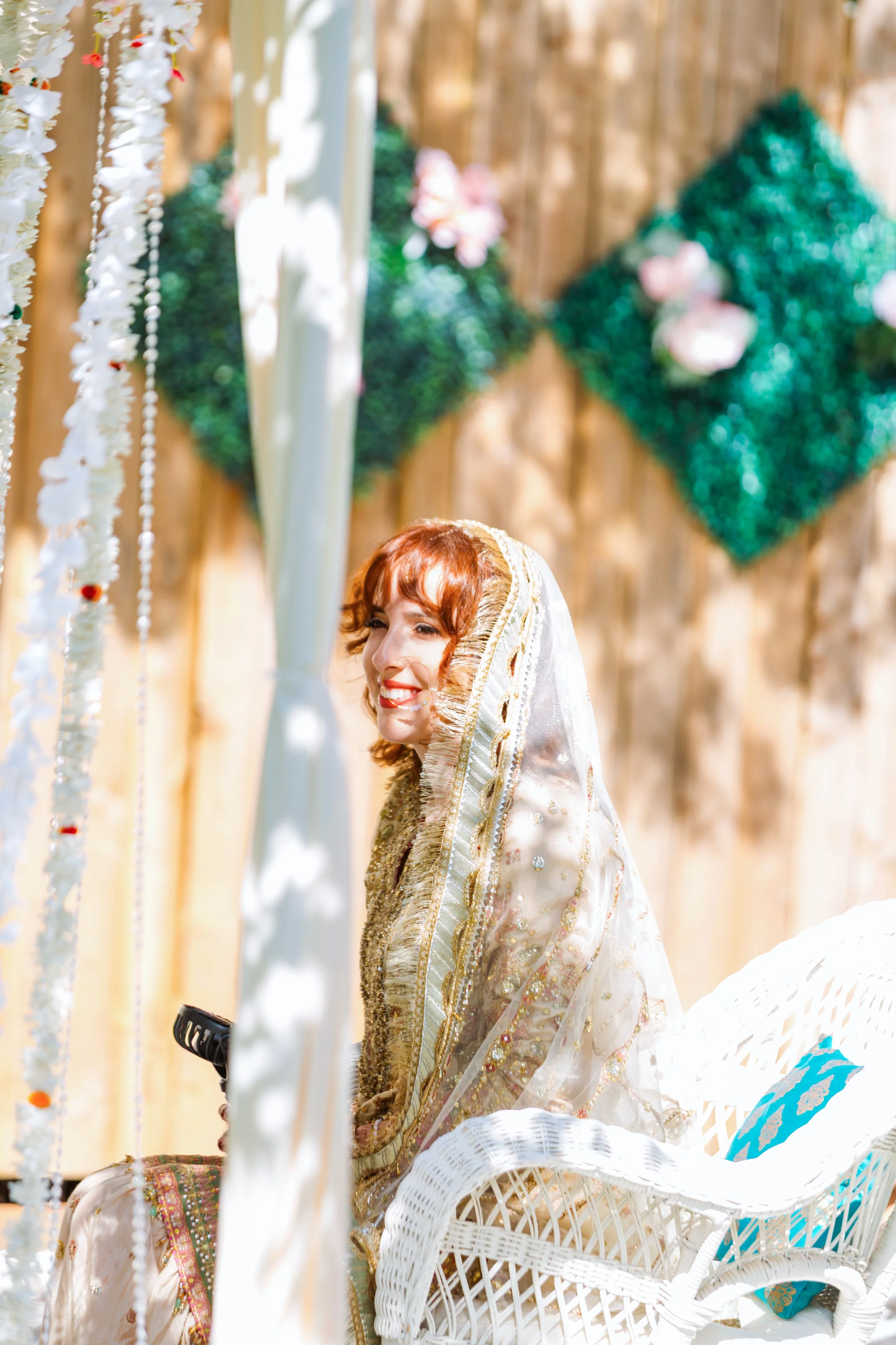 A woman with red hair smiling, dressed in an ornate cream and gold embroidered traditional outfit, sitting in a white wicker chair decorated with a matching veil, at an outdoor event with a wooden background and hanging floral and bead decorations.