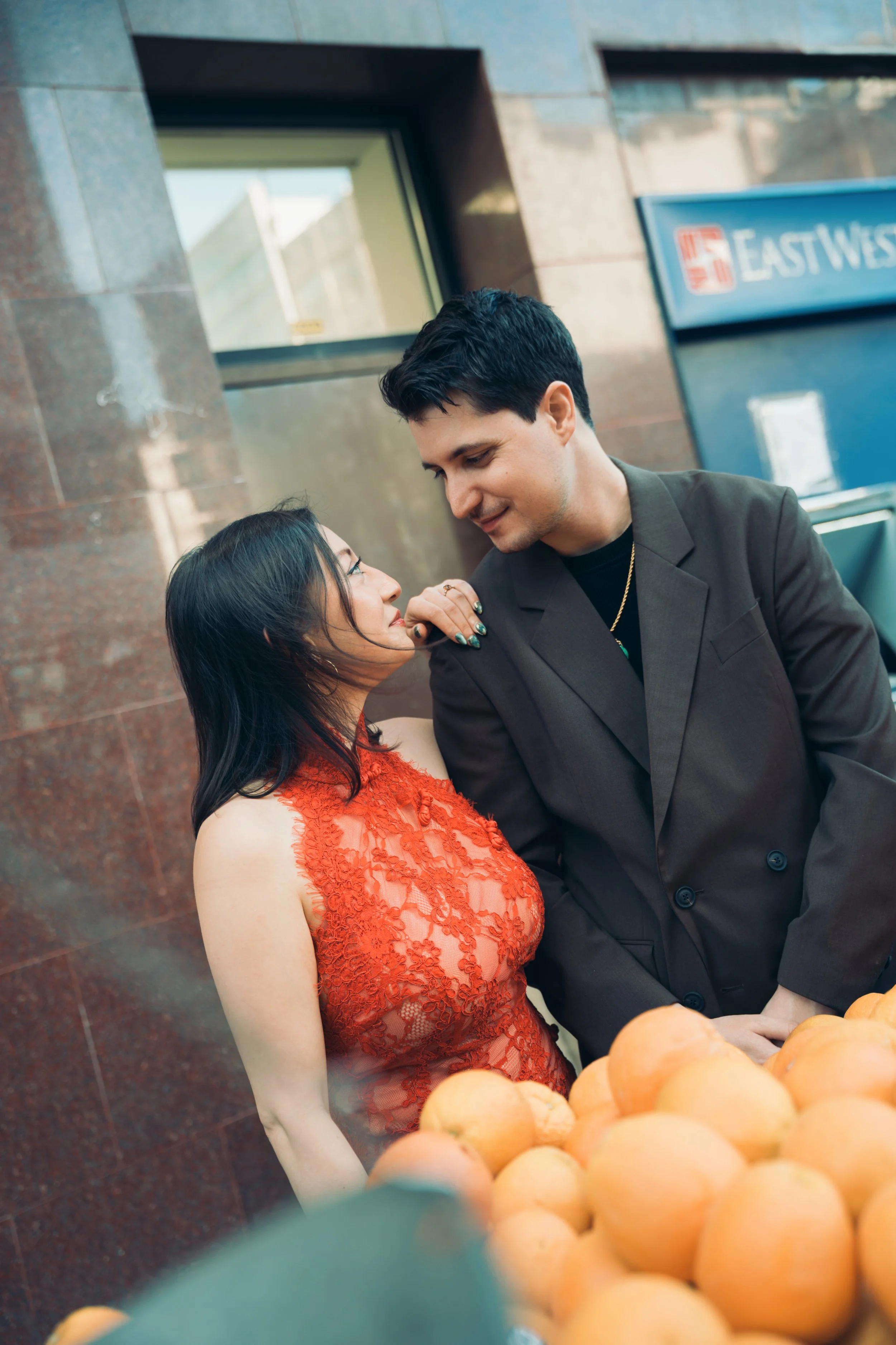 A man and woman looking at each other, standing close together outside a building with a sign that reads 'EAST WEST' behind them. The woman is wearing a red lace dress, and the man is wearing a dark suit. There are light-colored pumpkins in the foreg