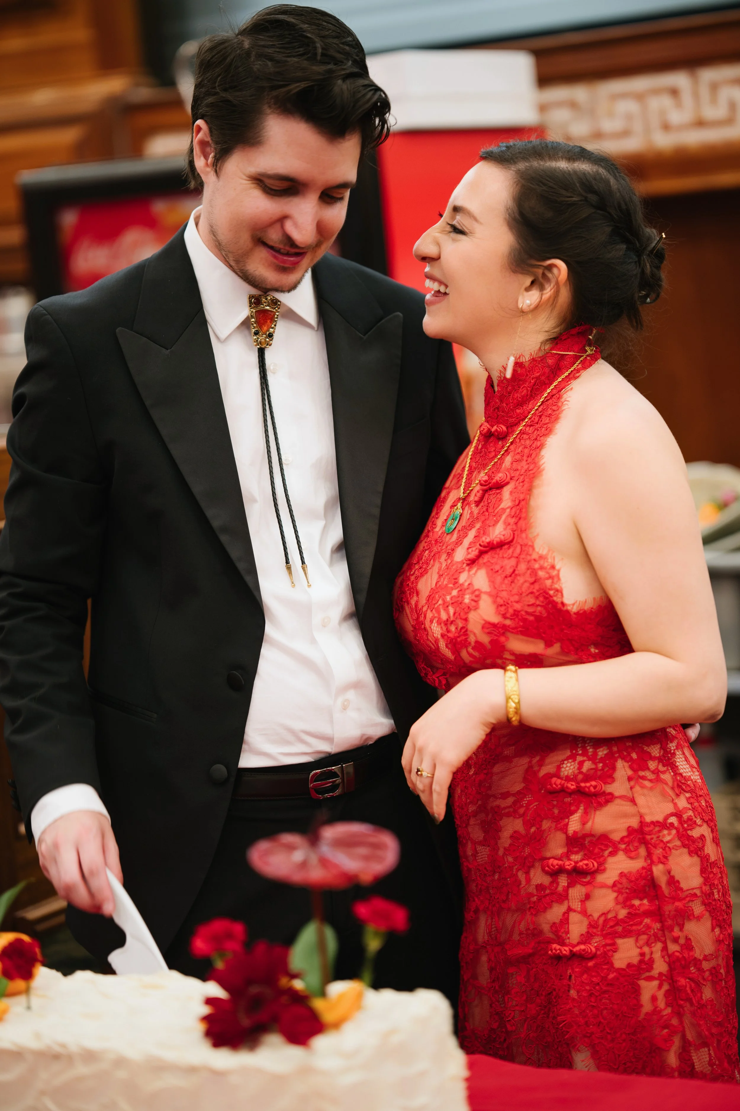 A man and woman dressed in formal attire are celebrating together, with the man cutting a cake and the woman smiling near him.