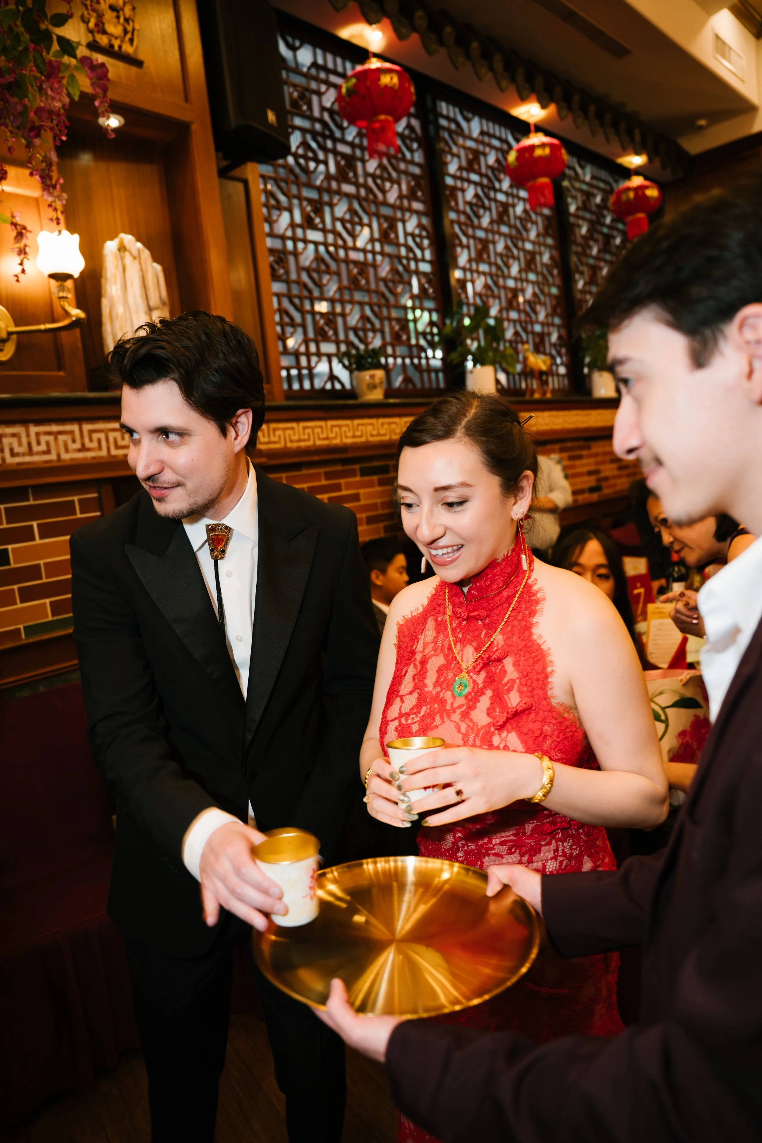 People participating in a traditional tea ceremony, with individuals dressed in formal attire, inside a restaurant decorated with red lanterns and Chinese-inspired decor.