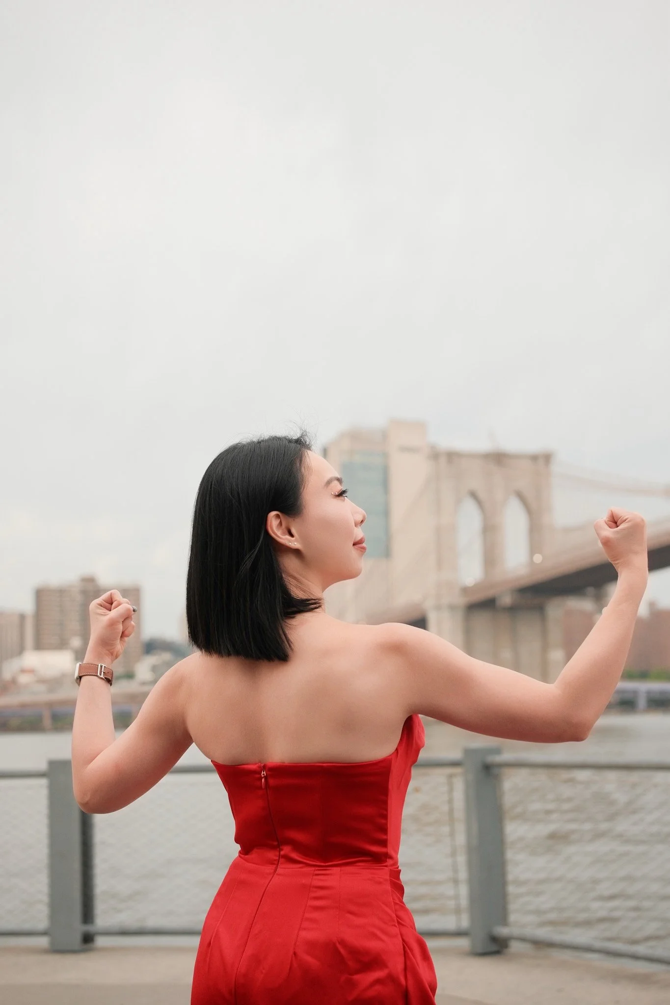 A woman in a red dress is standing outdoors on a bridge, flexing her arms, with the Brooklyn Bridge in the background.