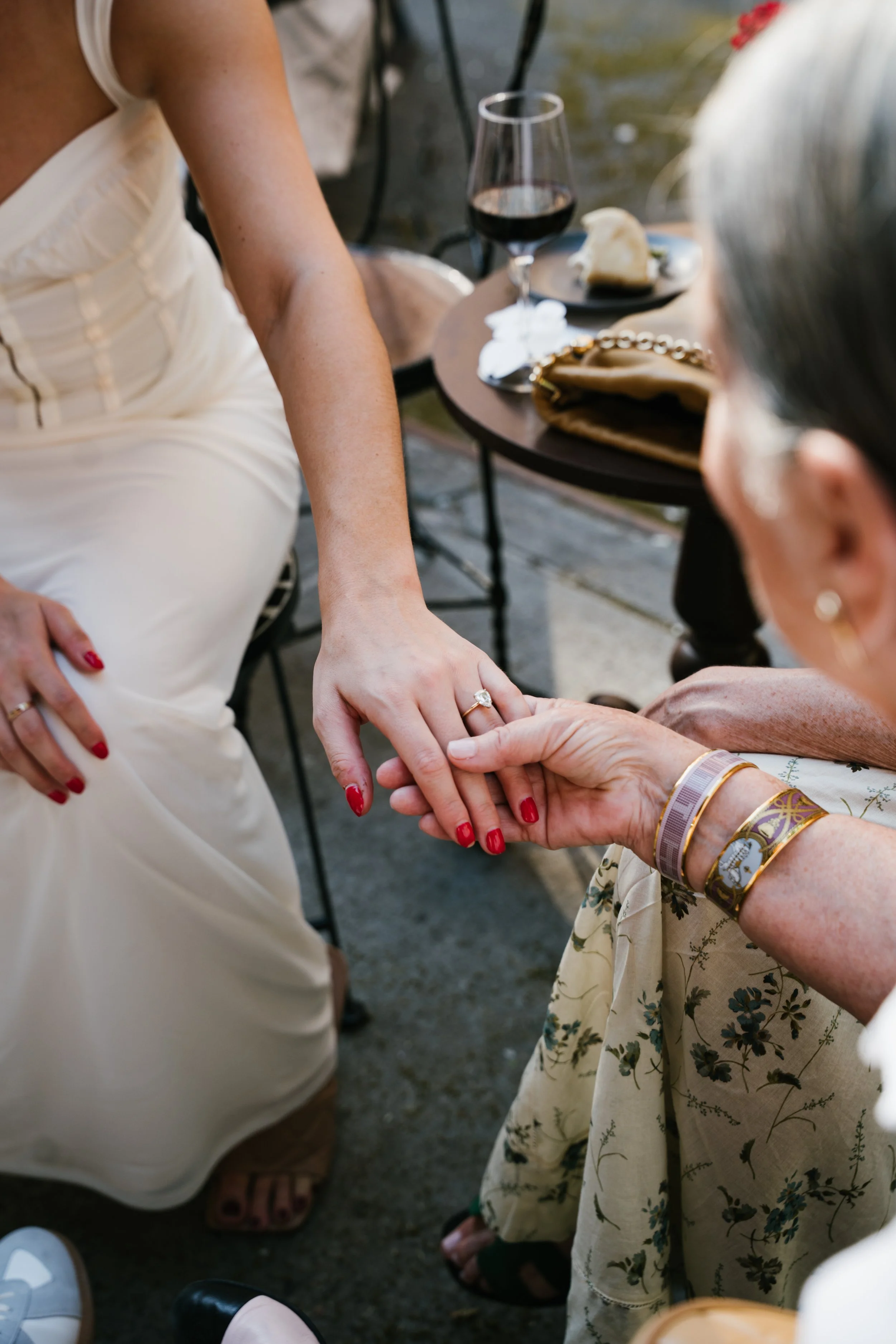 A woman in a white dress holding hands with an elderly woman in a floral dress, sharing a tender moment at an outdoor event with wine and dessert on a table nearby.