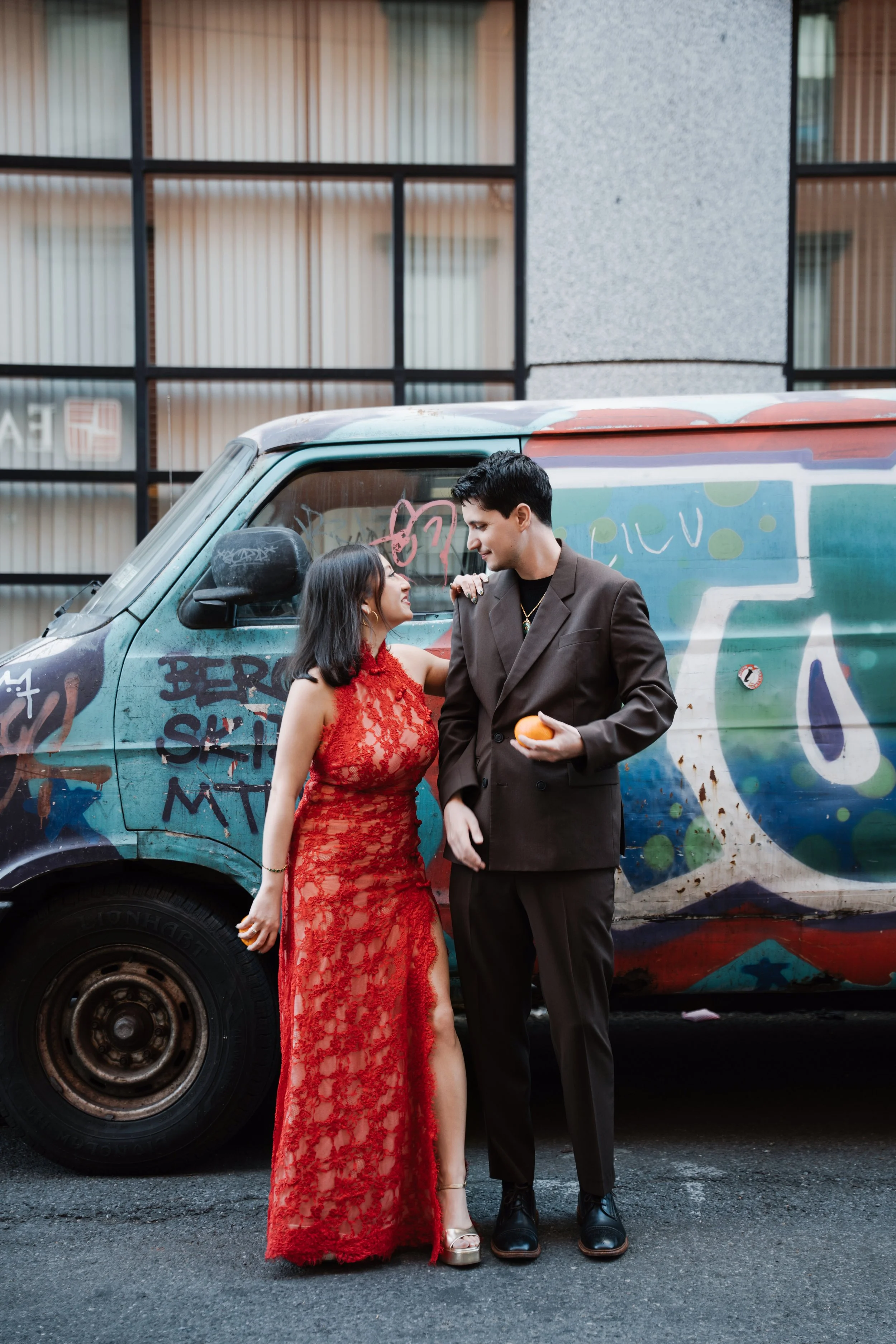 A man and woman standing beside each other in front of a graffiti-covered truck. The woman is wearing a red lace dress, and the man is dressed in a brown suit. They are looking at each other affectionately, with the man holding an orange. The backgro