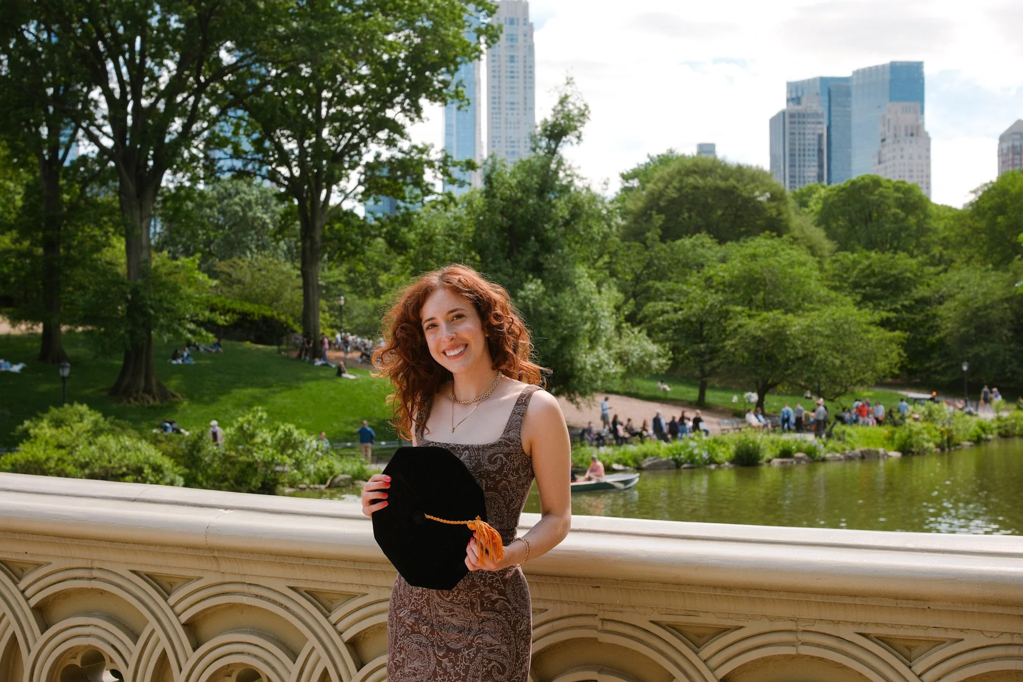 A smiling woman with curly red hair holding a black hat with an orange tassel, standing on a bridge over a river in a park with green trees and people in the background, with tall city buildings visible behind the trees.