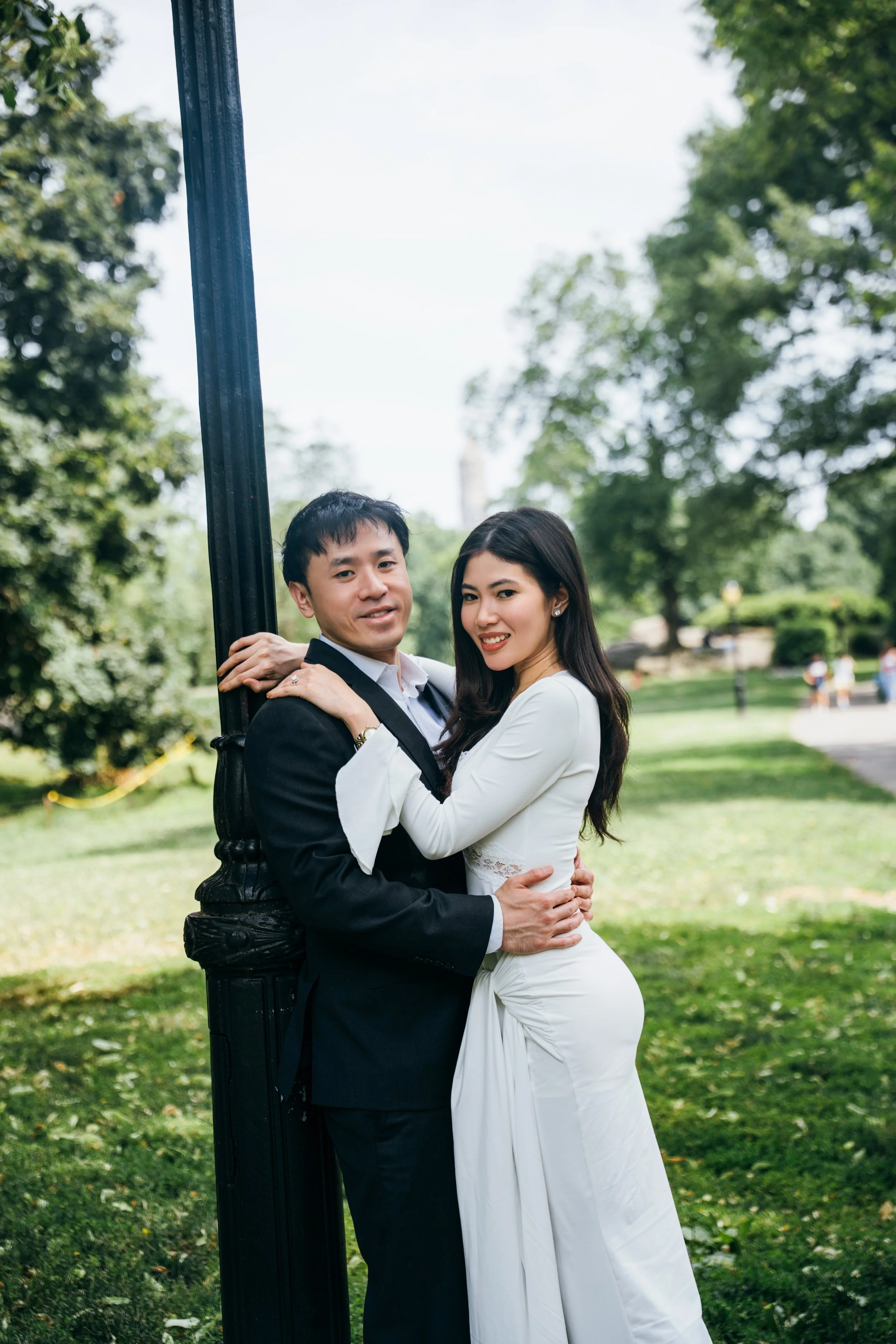 A couple dressed in wedding attire stands close together outdoors, holding each other and smiling. The man wears a black tuxedo with a white shirt, and the woman wears a white wedding dress with long sleeves and lace details. They are positioned next