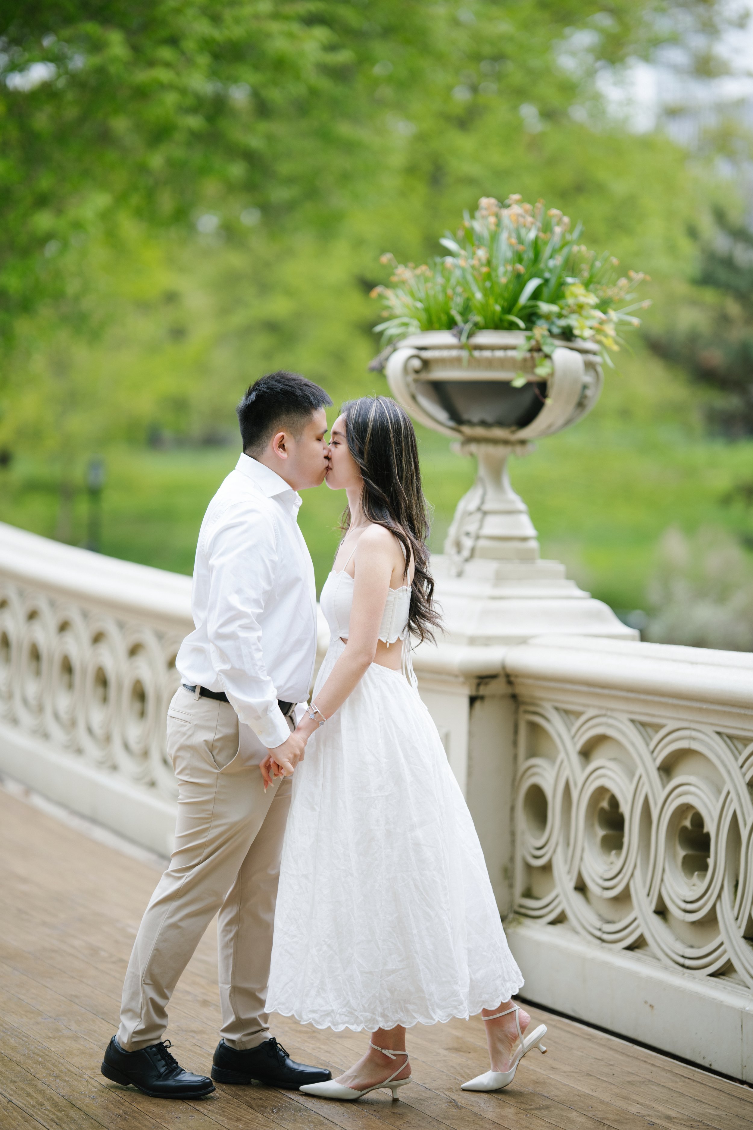 A couple holding hands and kissing on a bridge in a park, with a large decorative planter filled with green plants behind them.