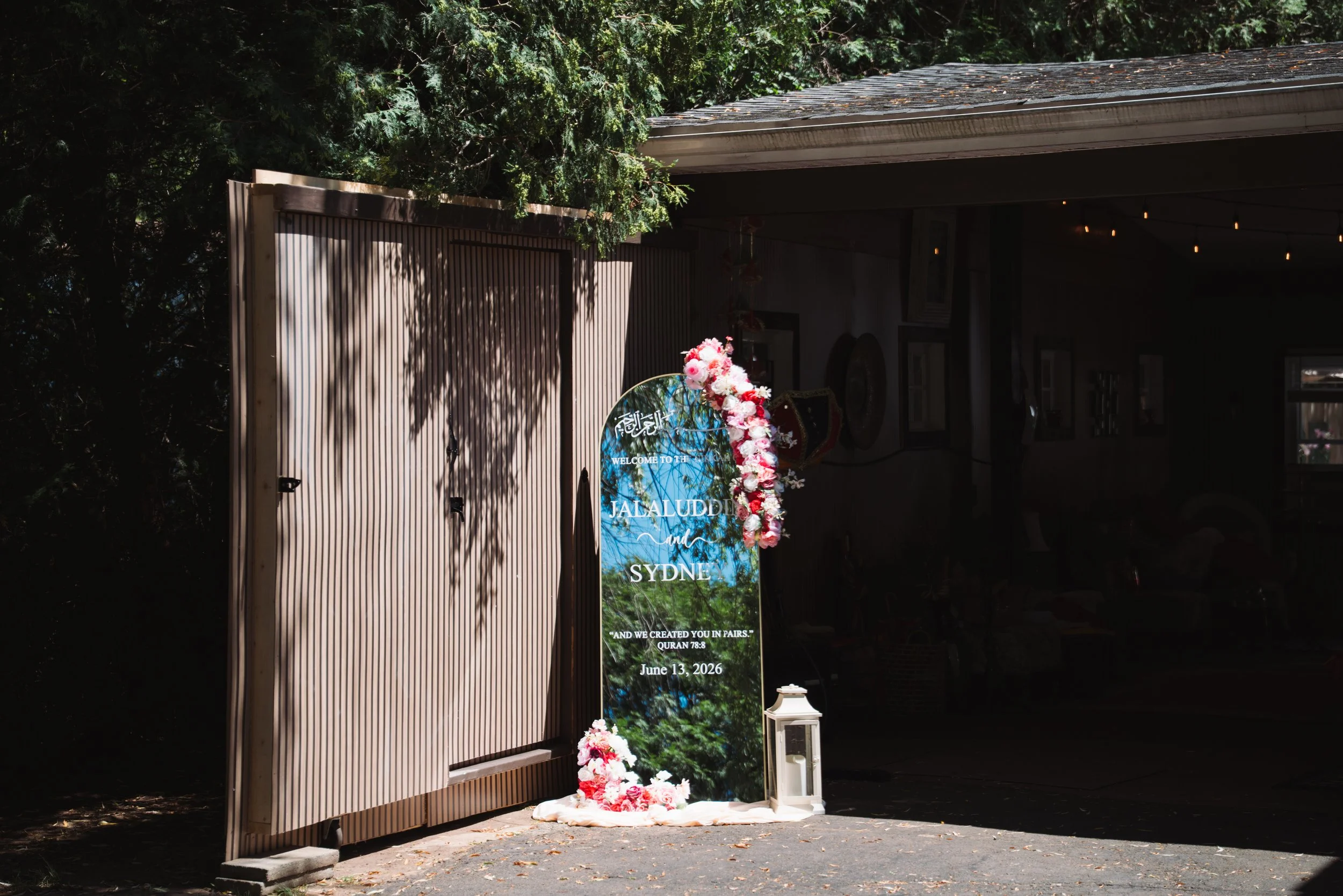 Decorative sign with floral arrangement for an event at Jalaluddin in Sydney, dated June 13, 2026, placed in front of a wooden fence with trees and greenery in the background.
