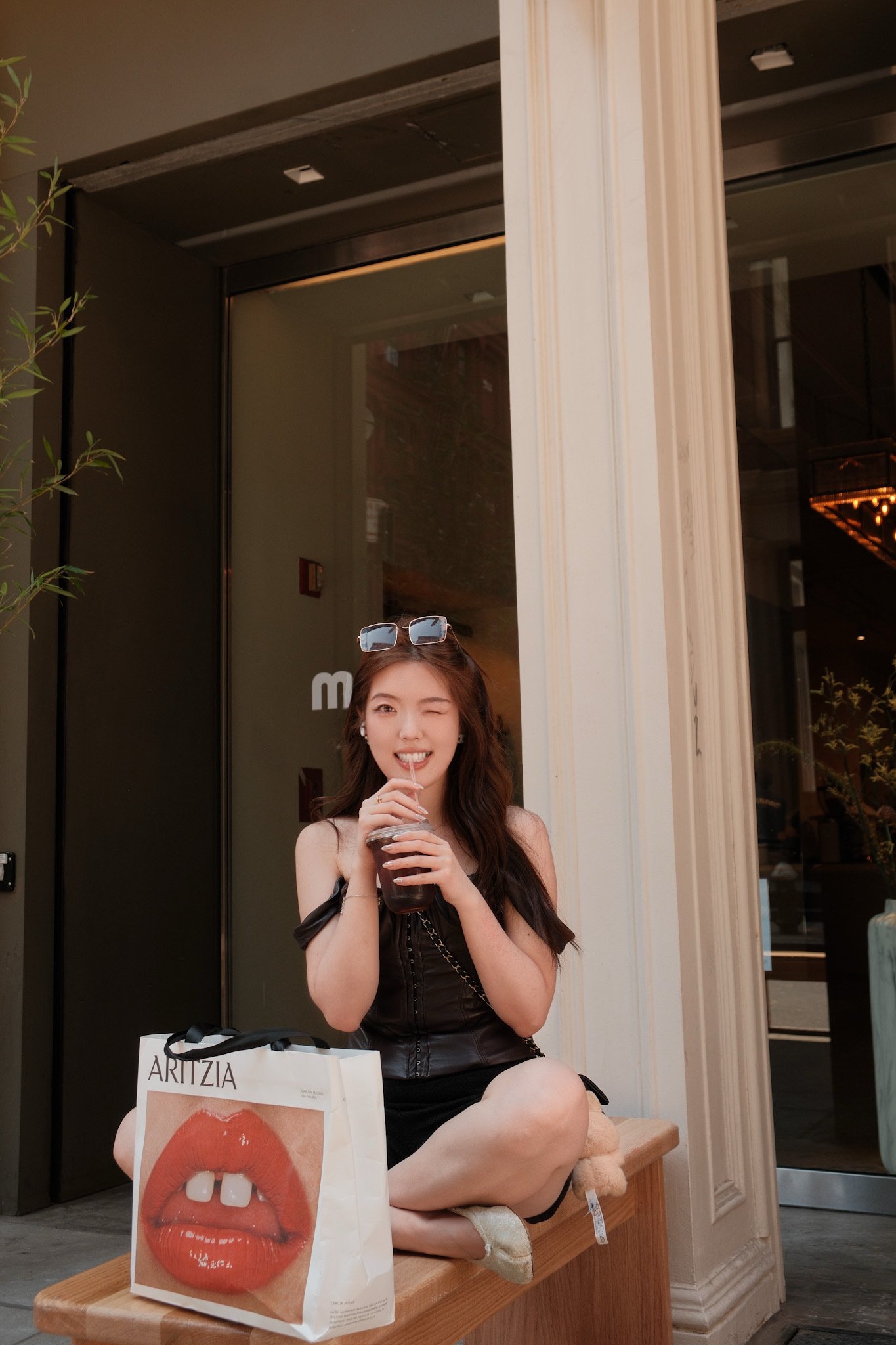 A young woman with long brown hair, wearing a black dress and sunglasses on her head, sitting on a wooden bench outside a building, drinking iced coffee with a straw, smiling and winking. In front of her, there is a shopping bag with a large image of