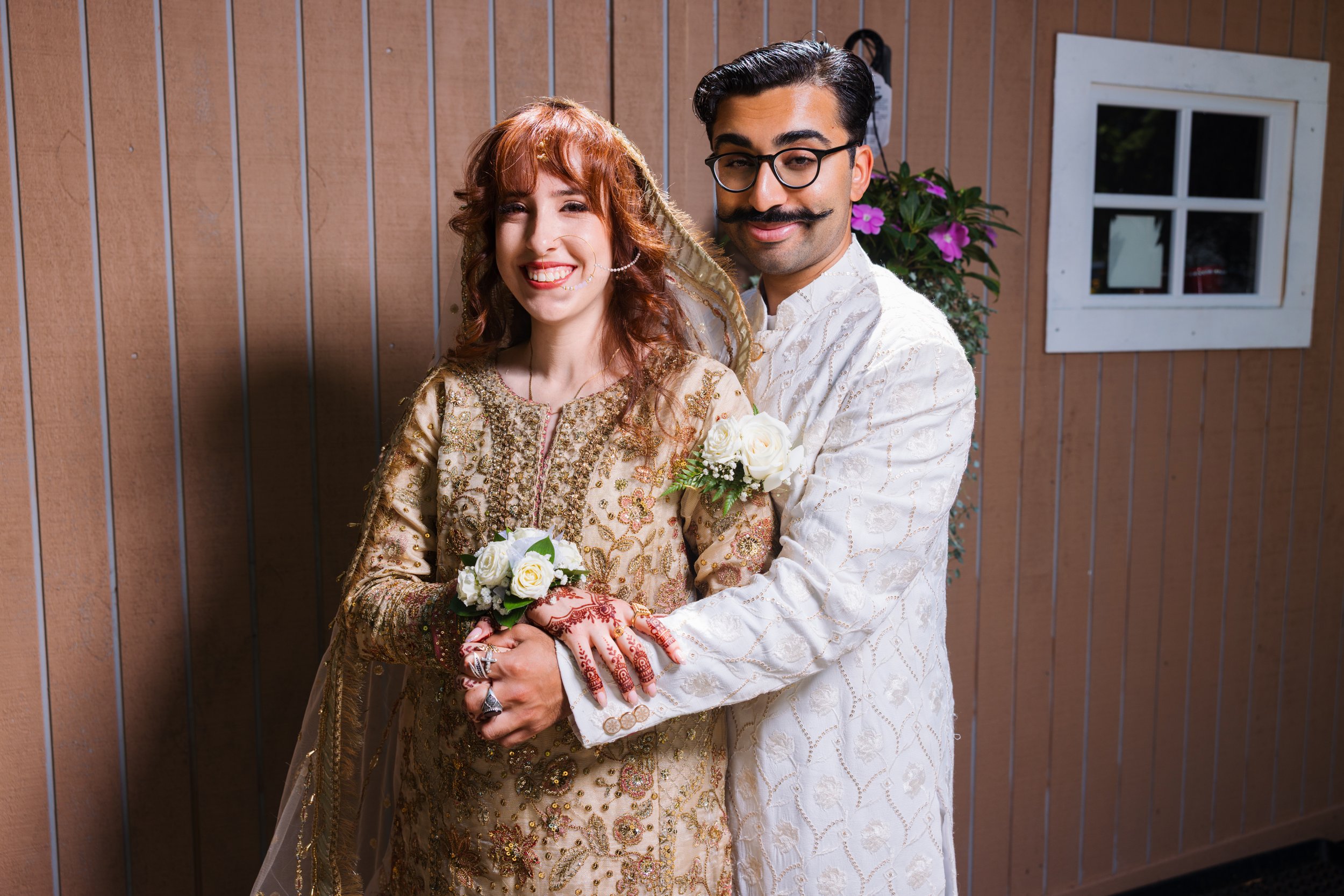 A wedding couple dressed in traditional attire, standing outdoors in front of a wooden wall with purple flowers and a window behind them, smiling happily.