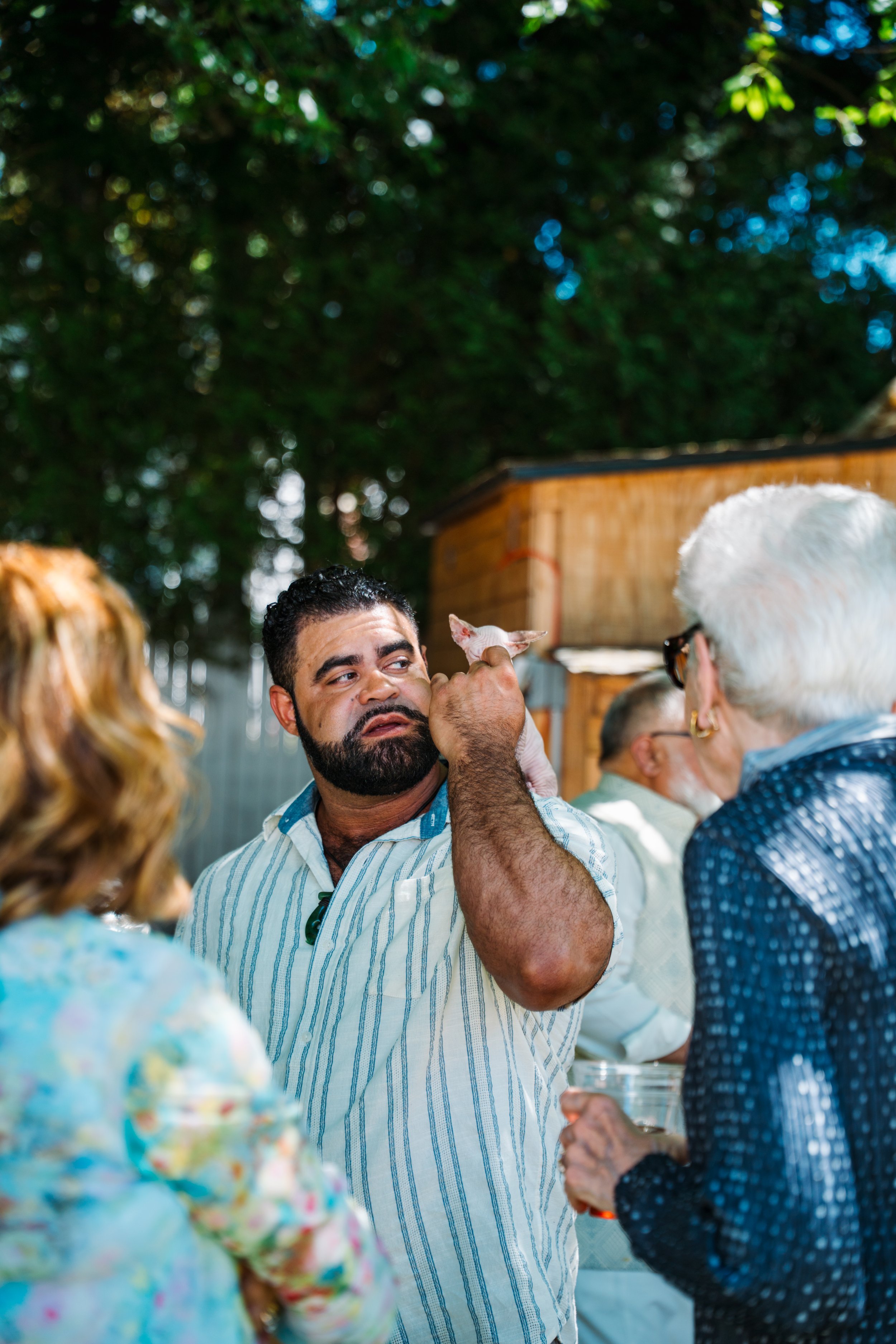A man with a beard and striped shirt holding a small dog, surrounded by people at an outdoor gathering.