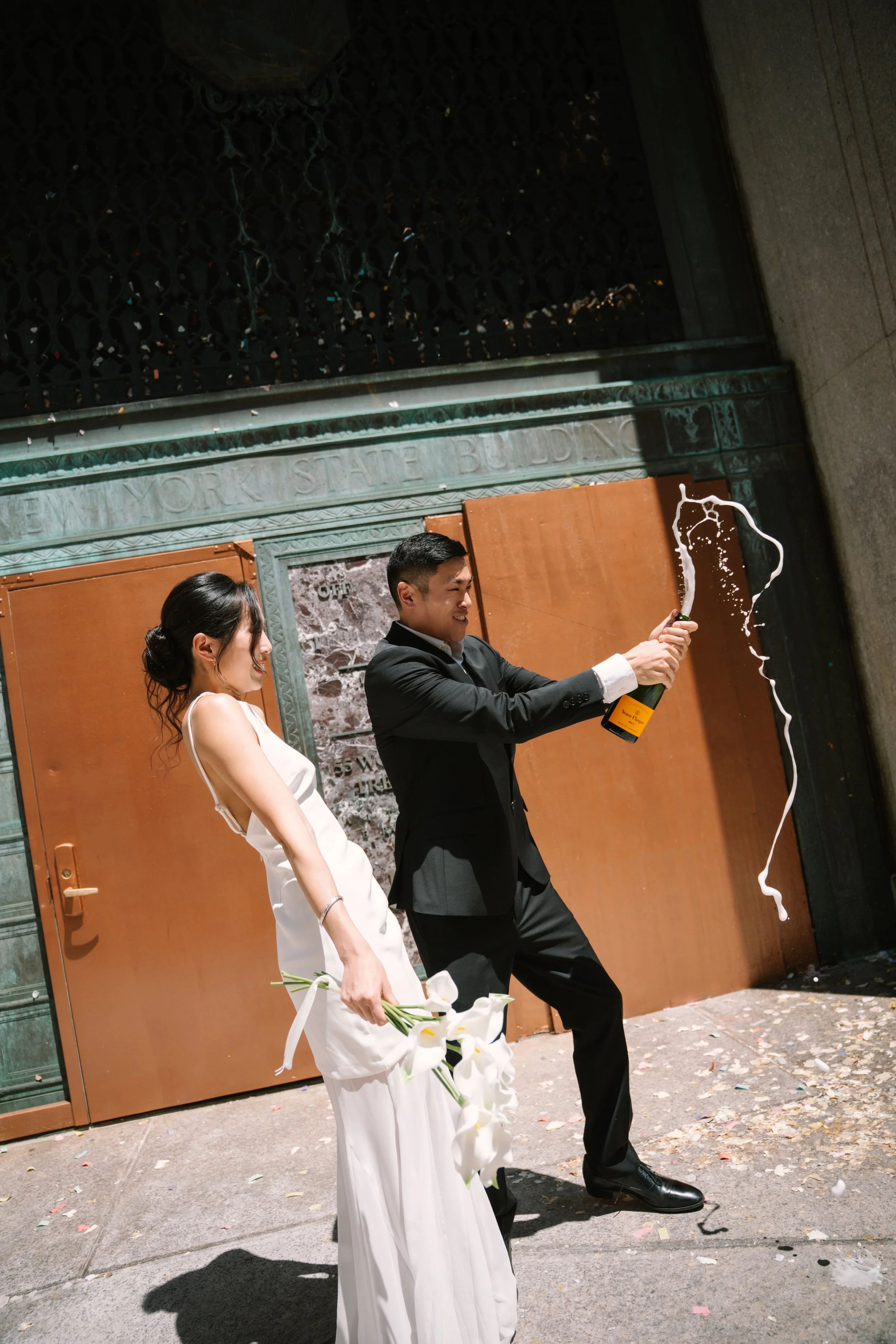 A bride and groom celebrating at their wedding outside at night. The groom is opening a champagne bottle, and foam is flying out. The bride is holding a bouquet of white flowers, and they are both dressed in wedding attire. There is confetti on the g