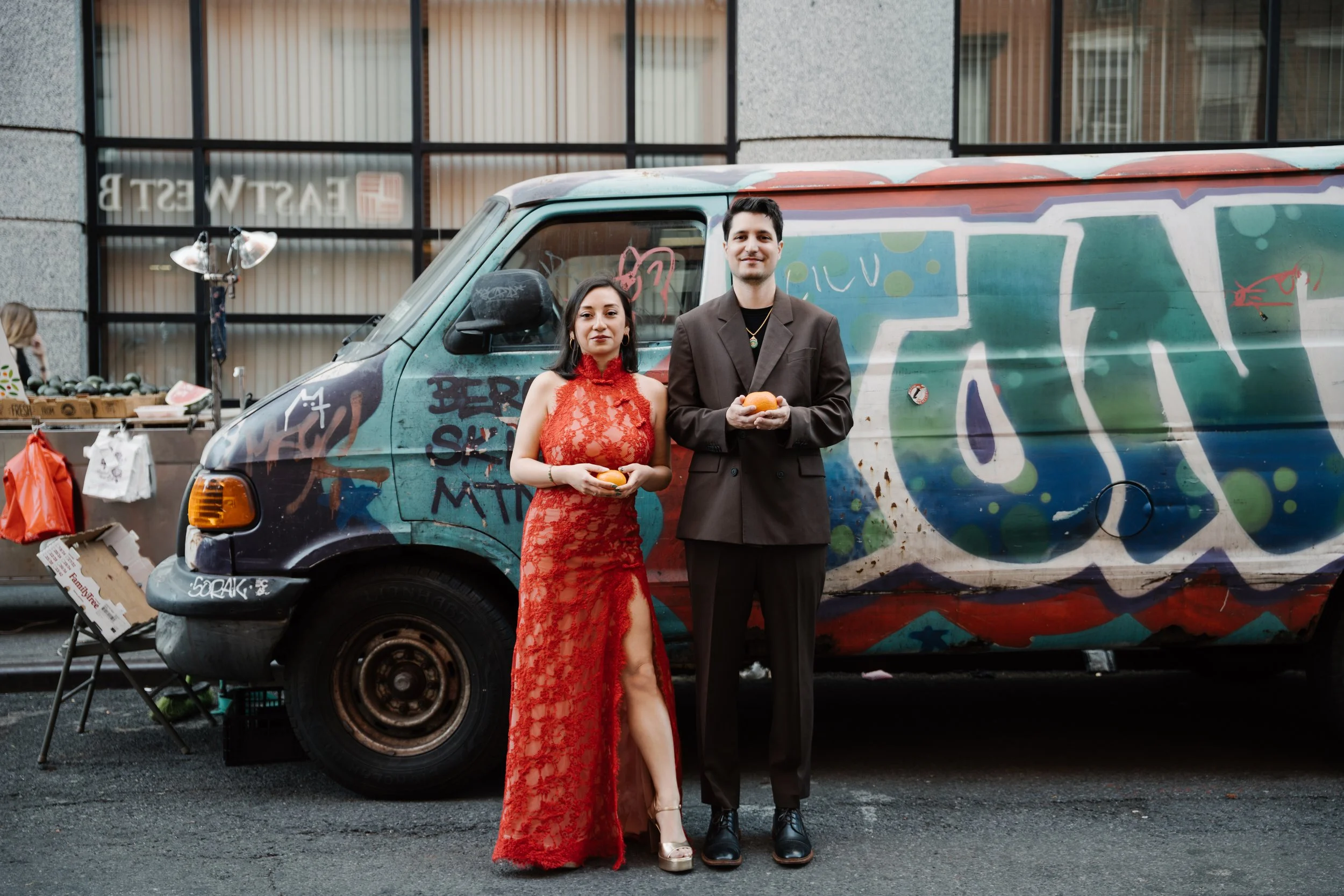 A man and woman stand side by side in front of a graffiti-covered van on a city street. The woman wears a red lace dress, and the man is dressed in a dark suit with a gold necklace. They are both holding small round objects.