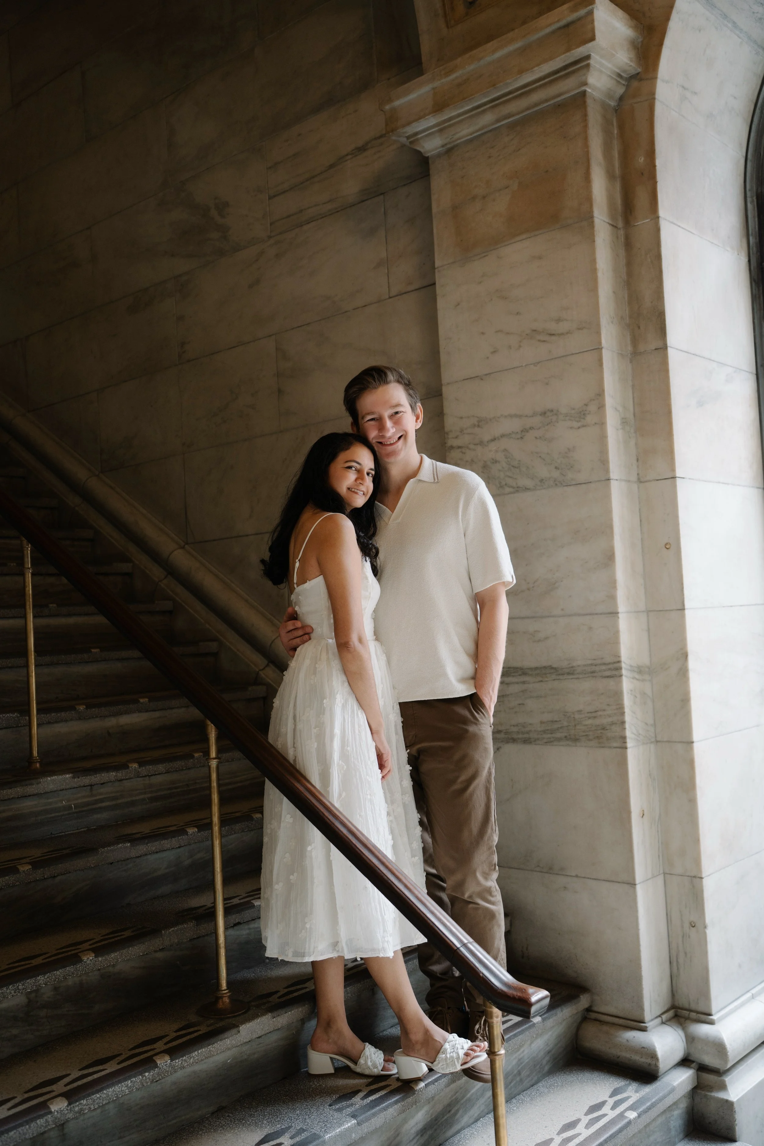 A smiling young woman wearing a white dress and white heeled shoes, standing on a staircase next to a smiling young man wearing a white shirt and beige pants in a marble building.