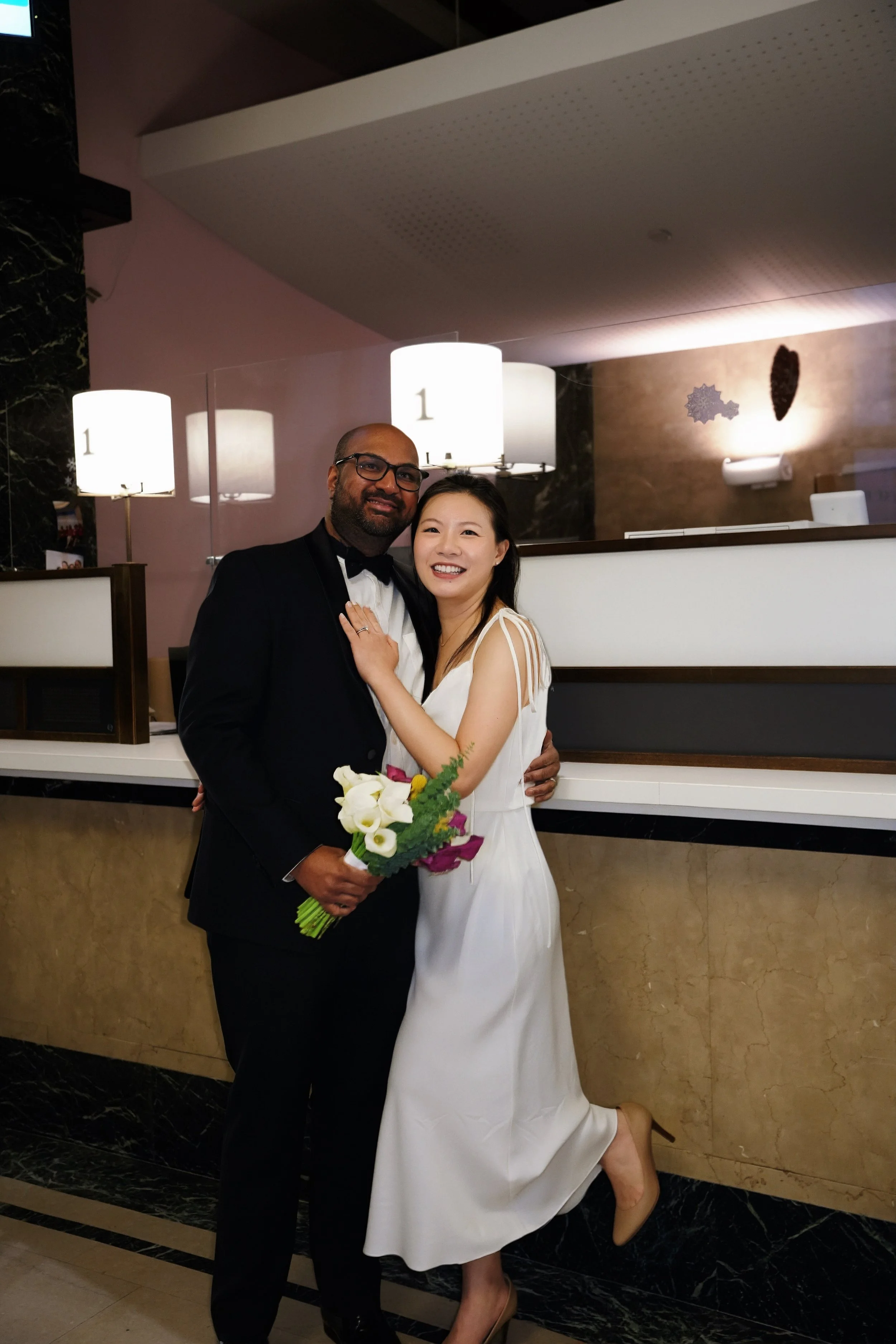 A happy couple dressed in wedding attire, posing for a photo in a hotel lobby. The man is wearing a black tuxedo with a bow tie, and the woman is in a white wedding dress, holding a bouquet of flowers. They are smiling and embracing each other.