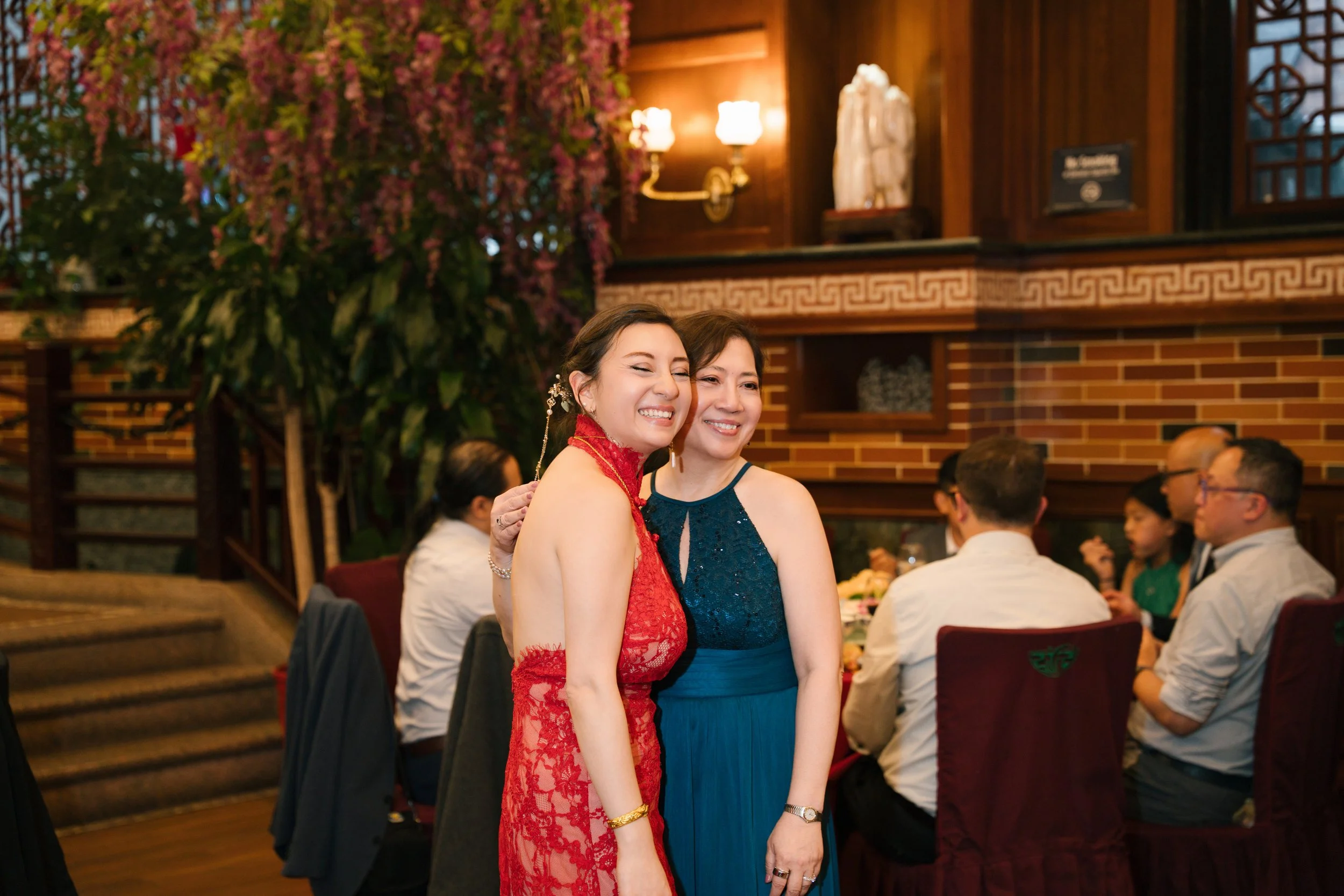 Two women smiling and hugging at a formal event, with other guests seated at tables in the background, indoors with wooden walls, decorative lighting, and floral arrangements.