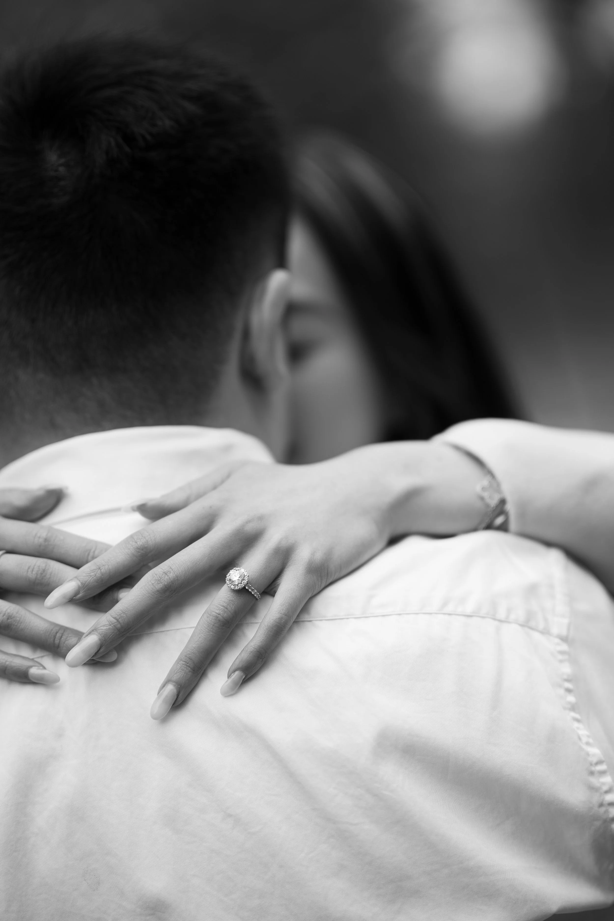 Close-up of a couple embracing, with the woman's hands showing an engagement ring on her finger, resting on a soft fabric surface.