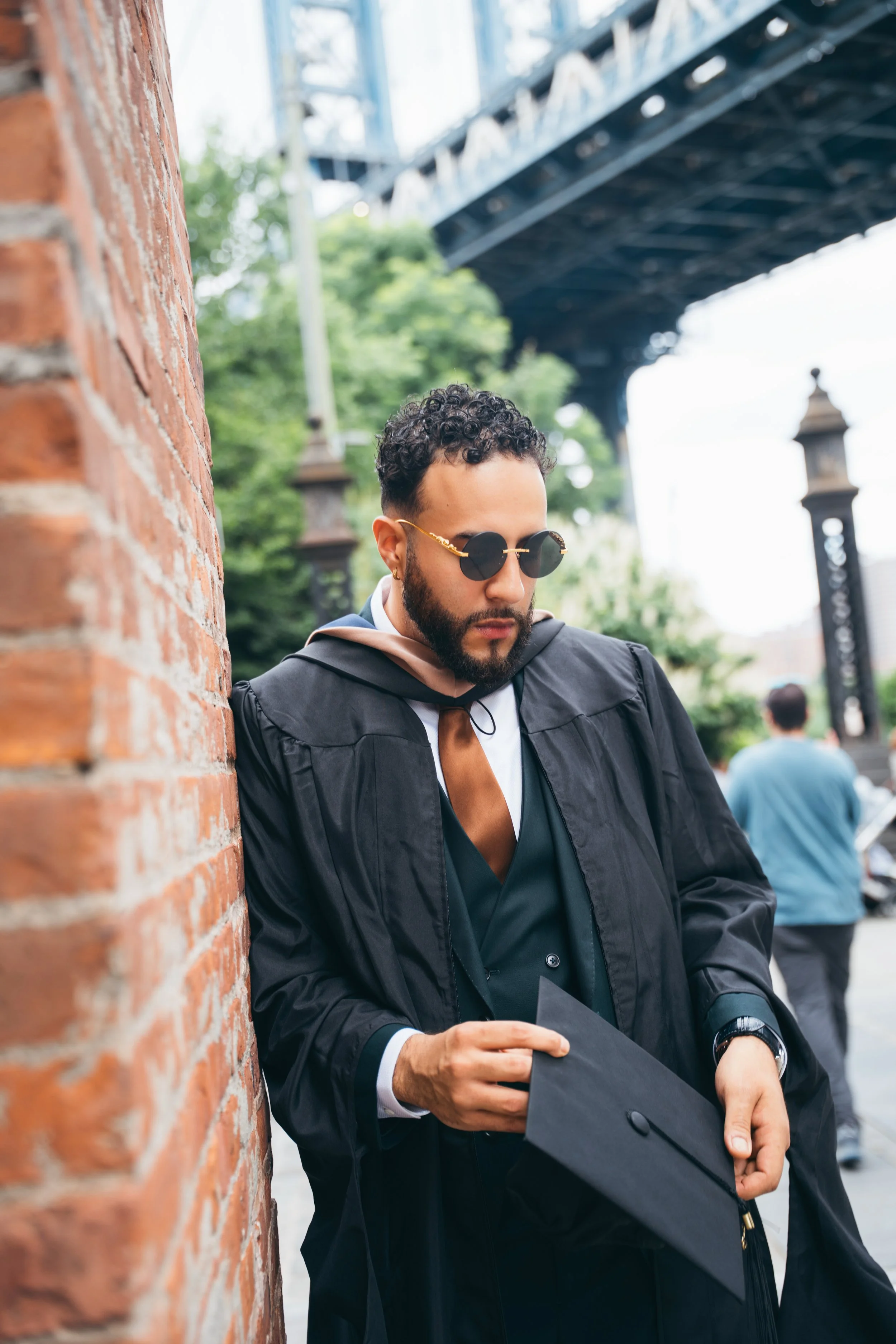 A young man with curly hair and sunglasses wearing a graduation gown and holding a diploma, leaning against a brick wall outdoors with a bridge and trees in the background.