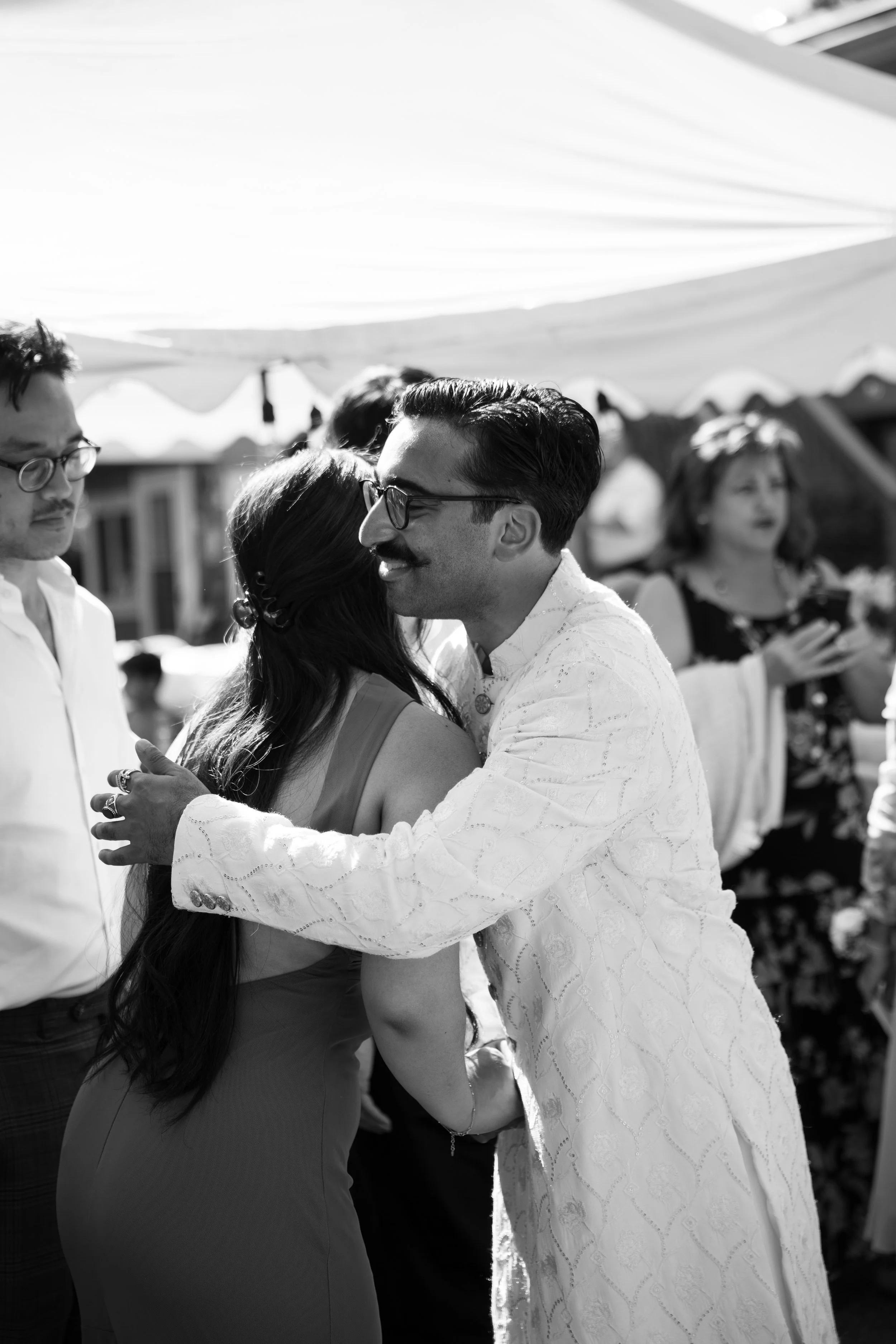 A man and woman wearing formal attire embracing and smiling at each other at an outdoor wedding or celebration, with other guests in the background under a tent.