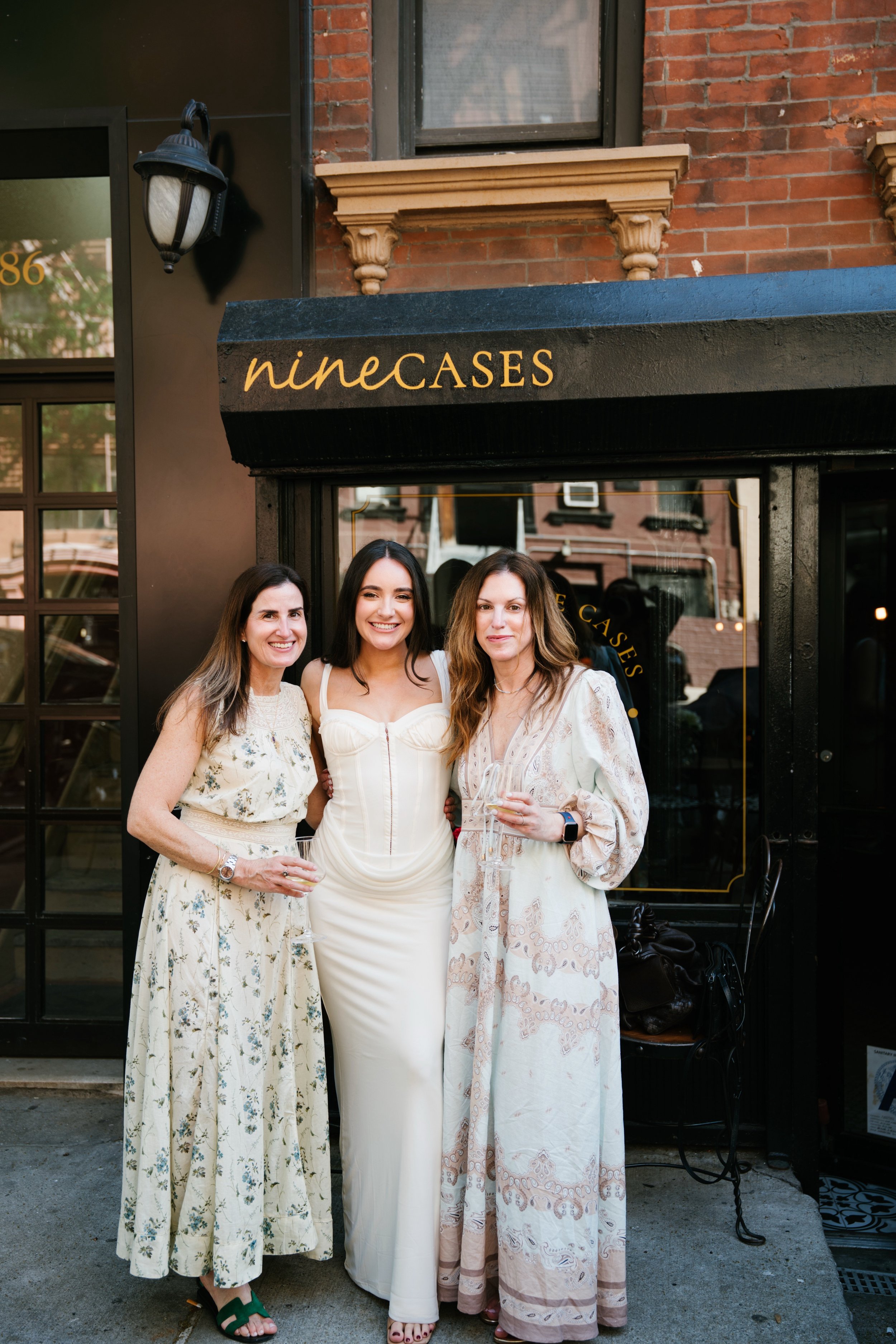 Three women standing together outdoors in front of a black storefront sign that reads 'nineCASES'. All are dressed in light-colored summer dresses and are holding glasses of white wine, smiling at the camera.