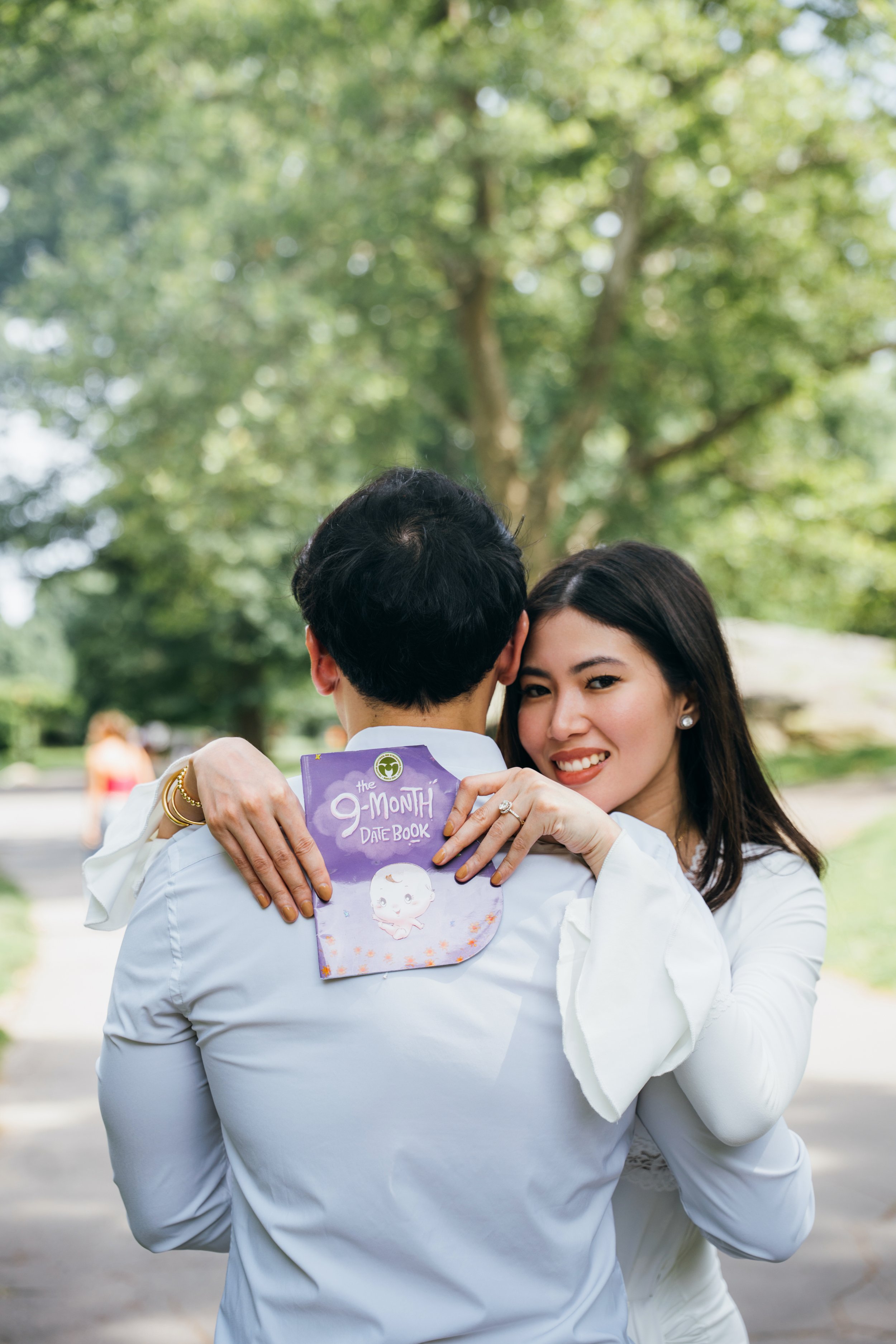 A woman smiling as she hugs a man, holding a purple baby memory book titled "The 9-Month Date Book" in an outdoor park setting with trees in the background.