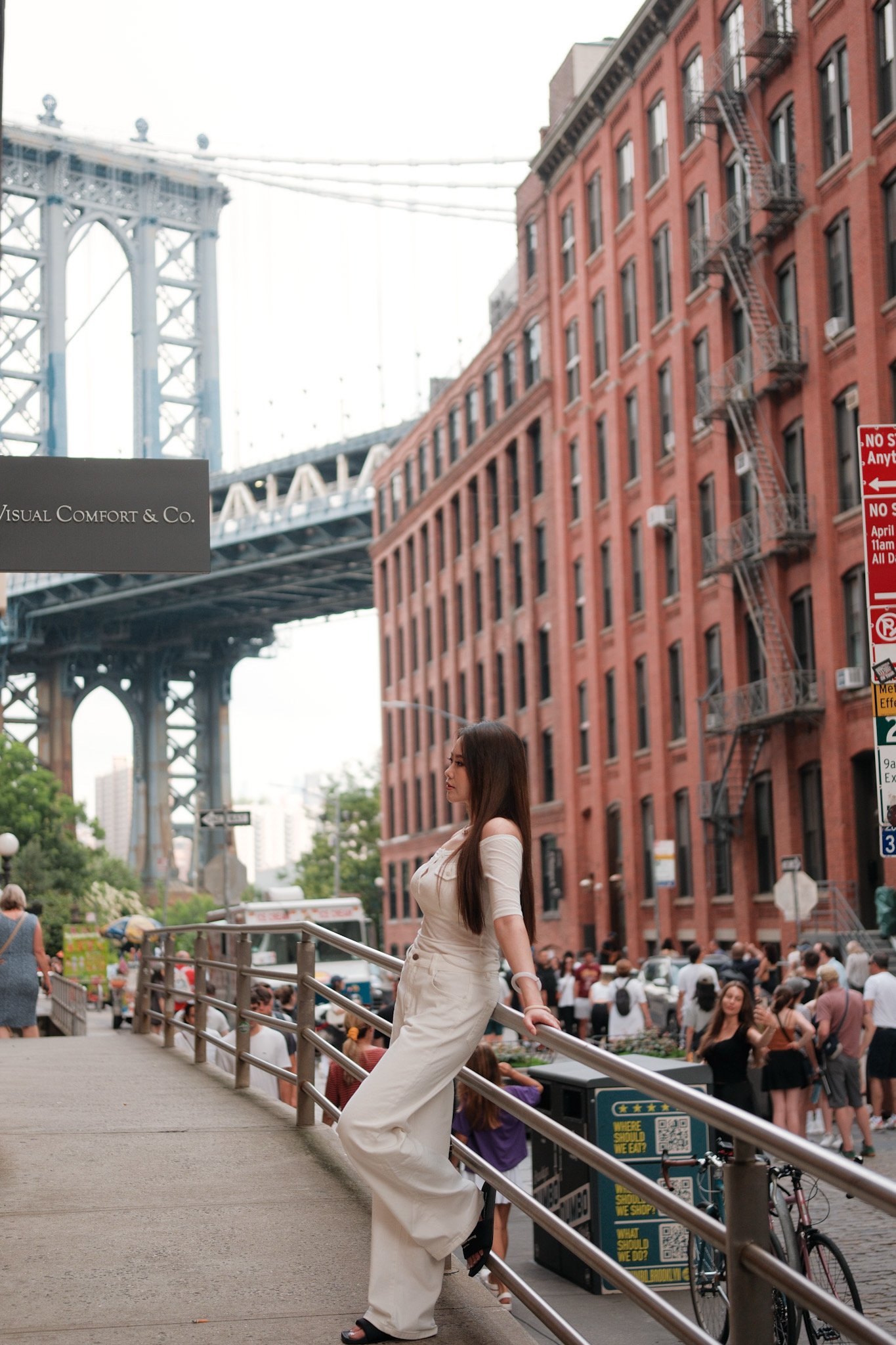 A woman in a white dress leaning against a fence in an urban outdoor setting with a bridge in the background; a crowd is visible behind her near a red brick building with fire escapes.