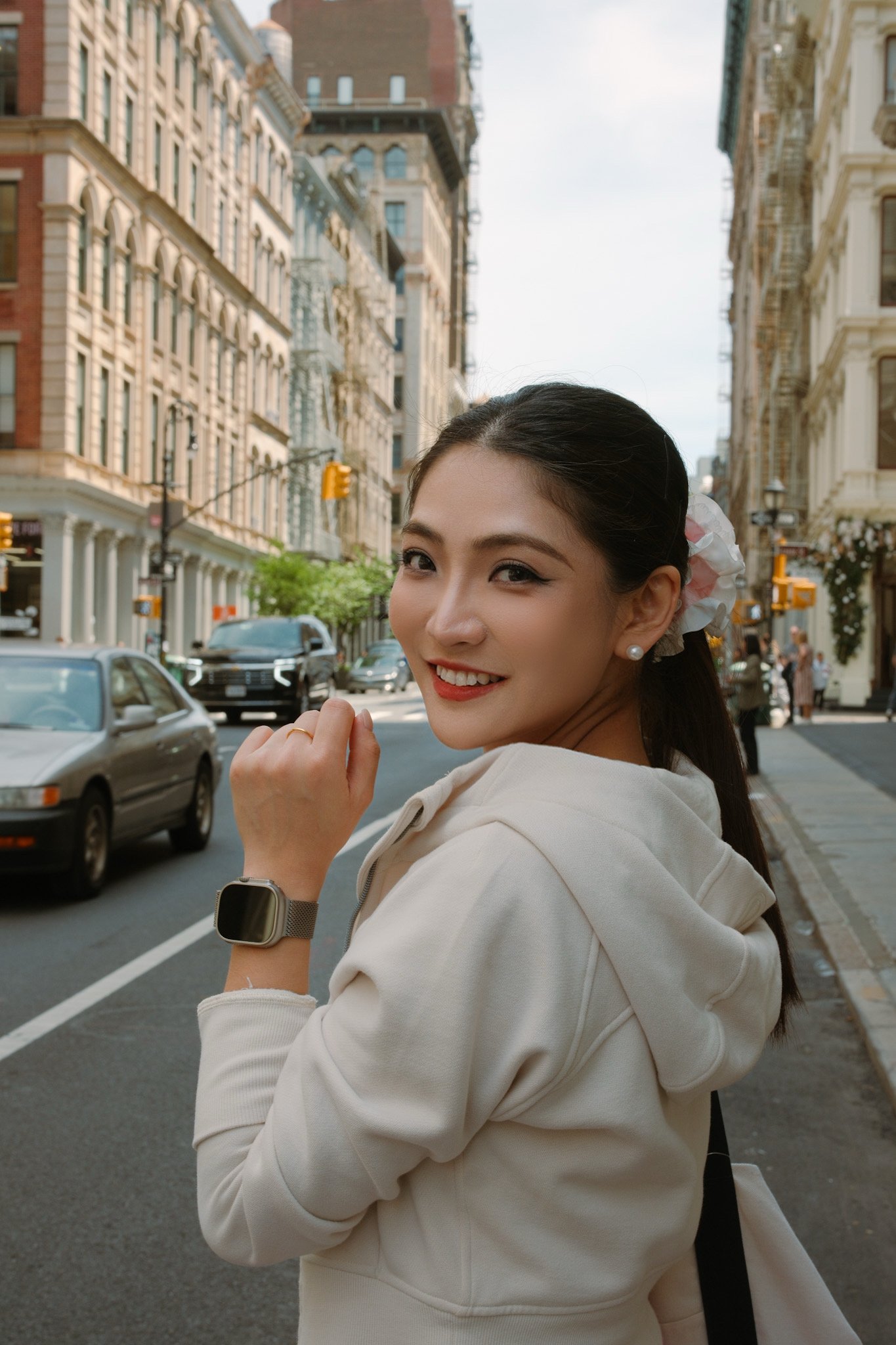 A young woman with dark hair tied back, wearing a beige hoodie and a smartwatch, smiling and looking over her shoulder while standing on city street with tall buildings, cars, and pedestrians.