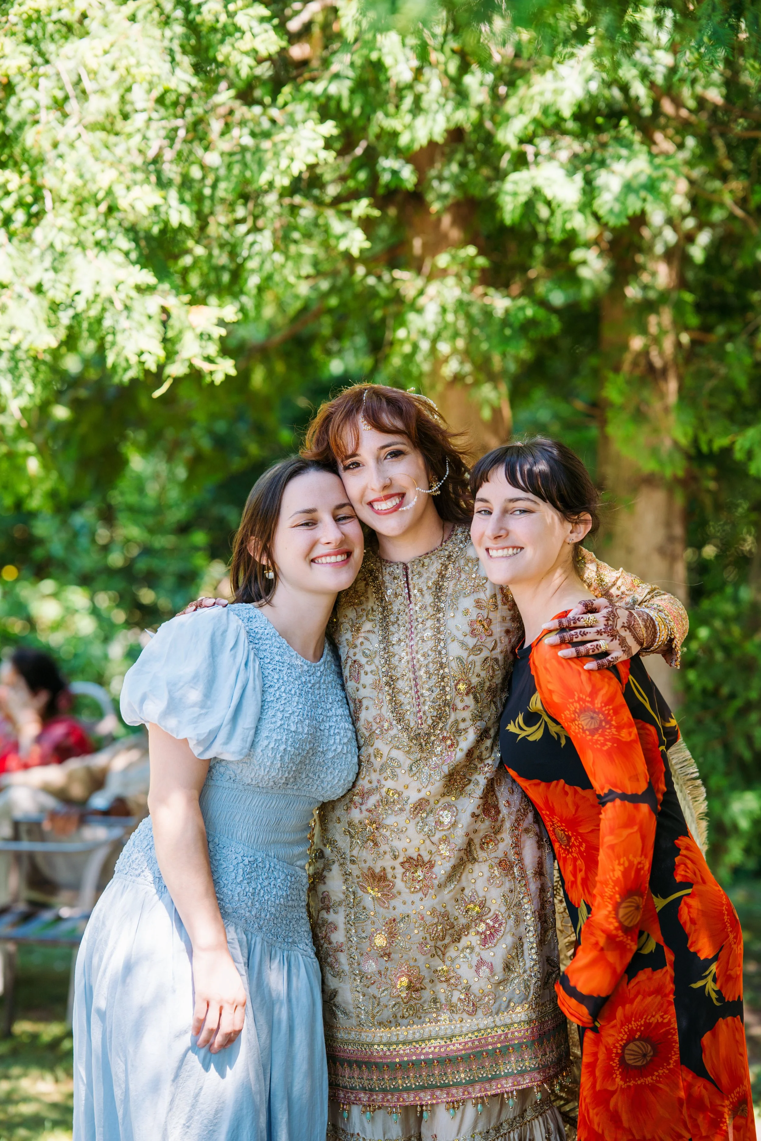 Three women standing close together outdoors under a large leafy tree, smiling at the camera. The woman in the center wears traditional South Asian attire, while the women on either side wear casual dresses.