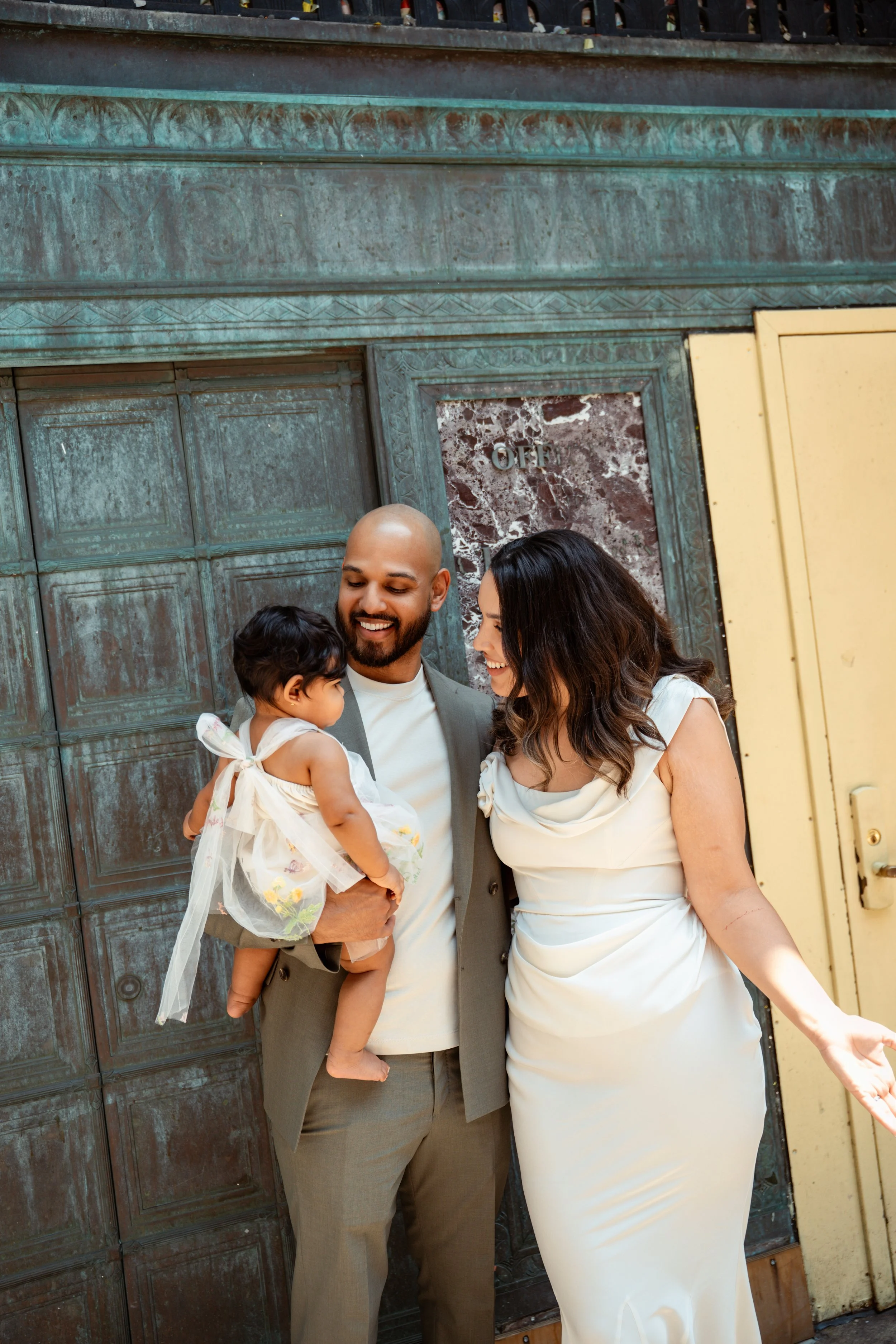 A happy family standing outside in front of a large, rustic metal gate. The man is holding a young girl, and the woman is smiling at the girl. The family appears to be celebrating a special moment together.