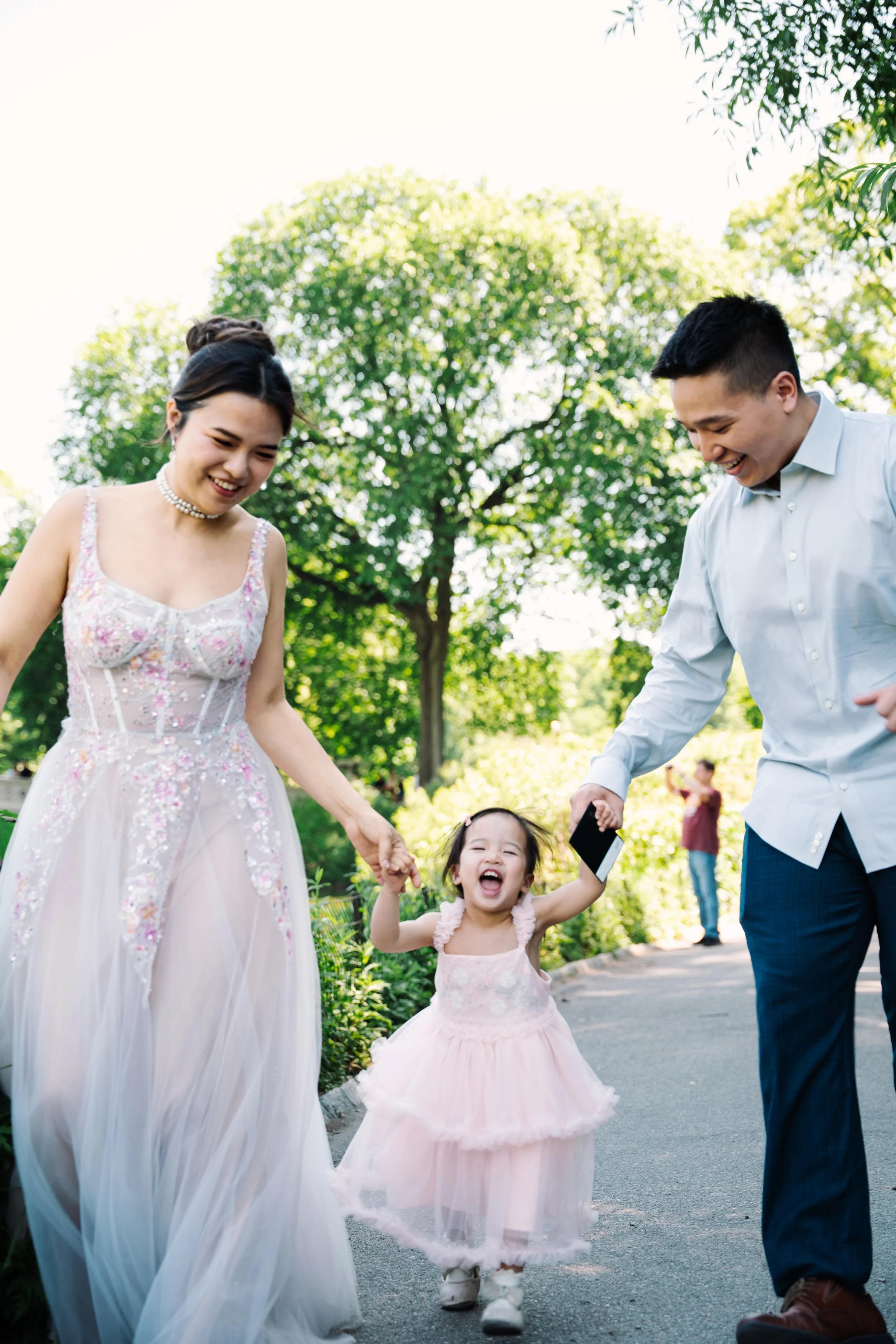 A family of three, including a woman in a light pink dress with floral embellishments, a man in a white shirt, and a young girl in a matching pink dress, are walking outdoors on a sunny day, holding hands and smiling.