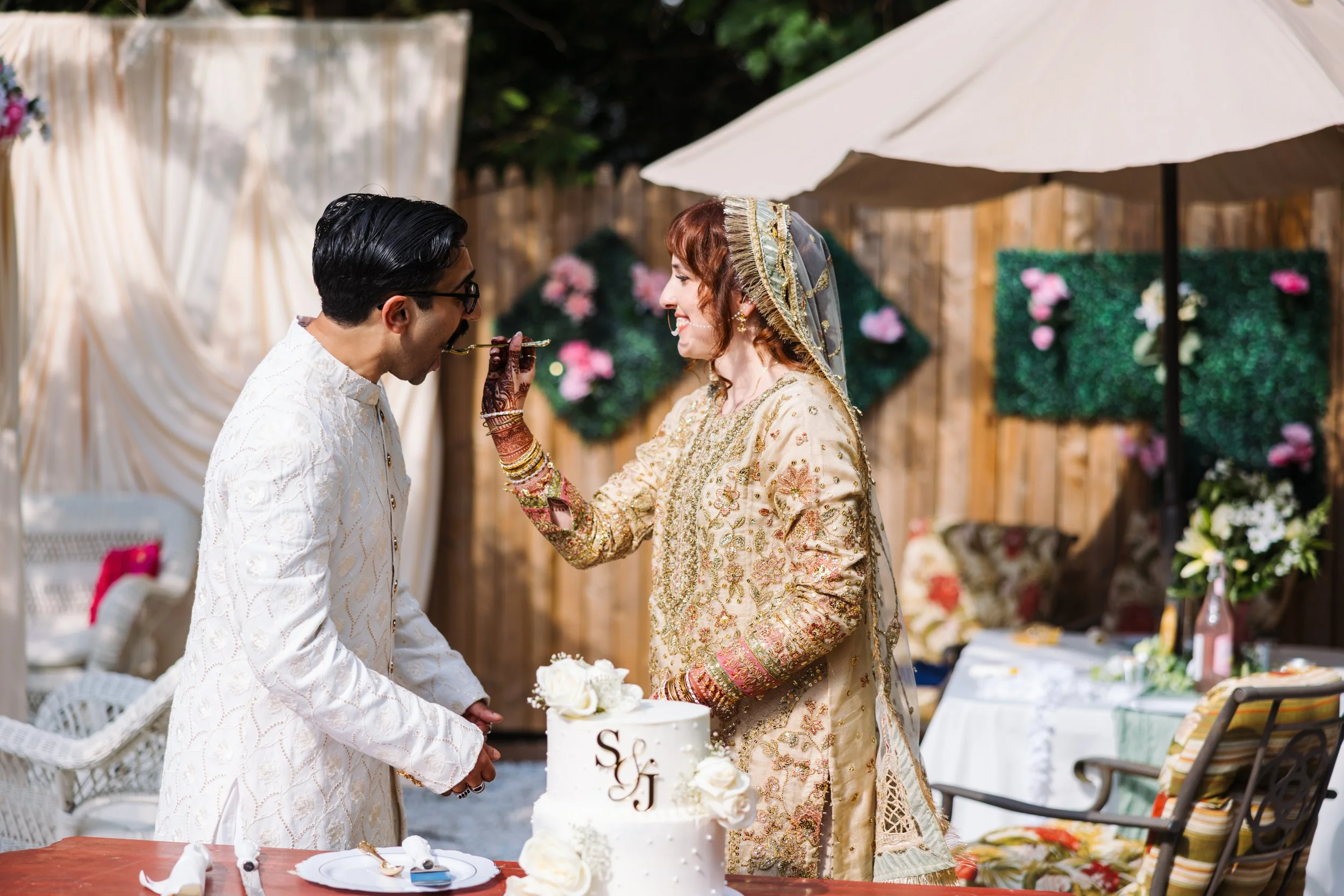 A bride and groom at their wedding, feeding each other cake outdoors surrounded by flowers and decorations.