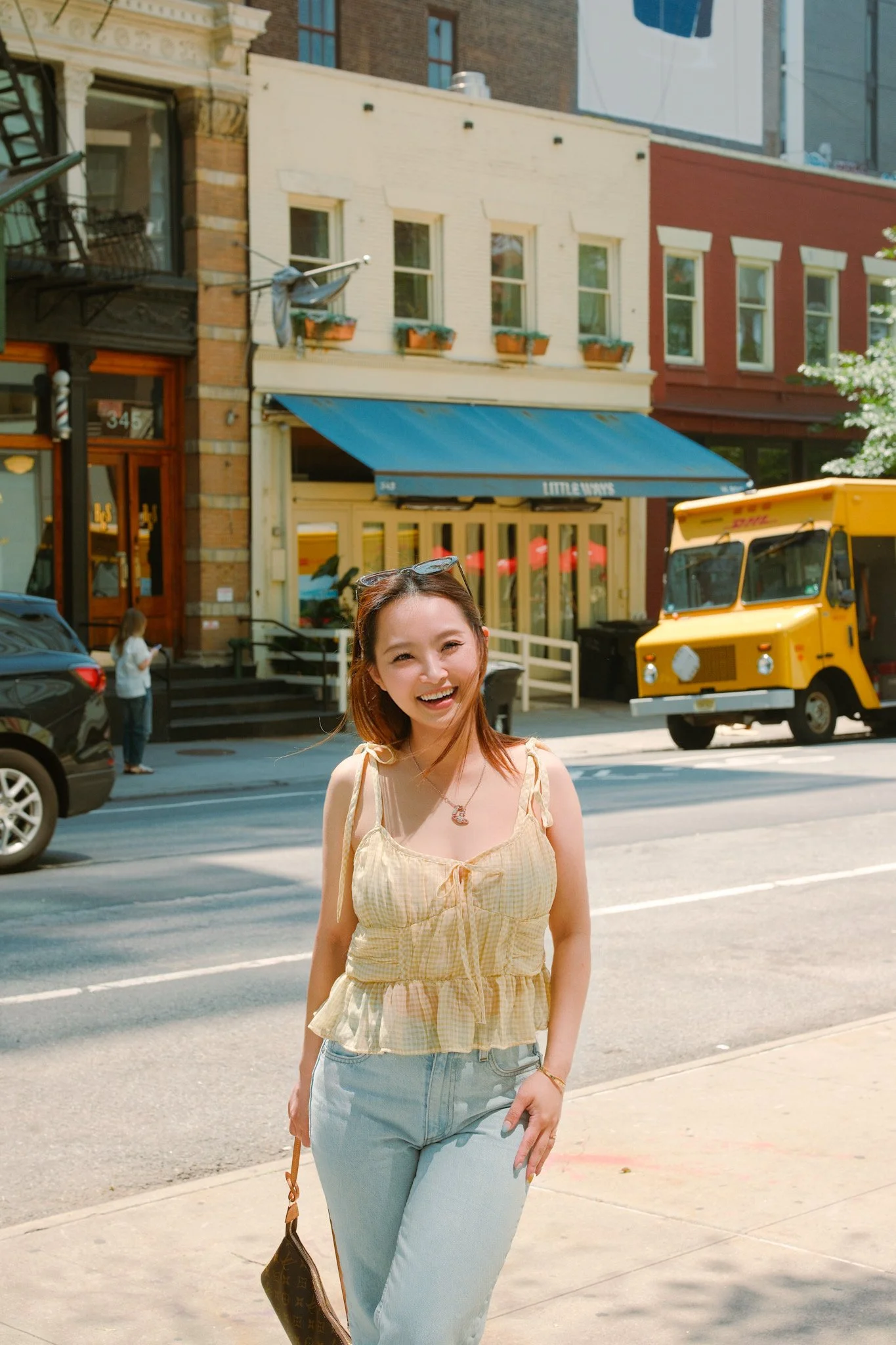 A smiling woman with light skin, wearing a yellow checked top and light blue jeans, walking on the street in front of buildings with storefronts and parked cars.