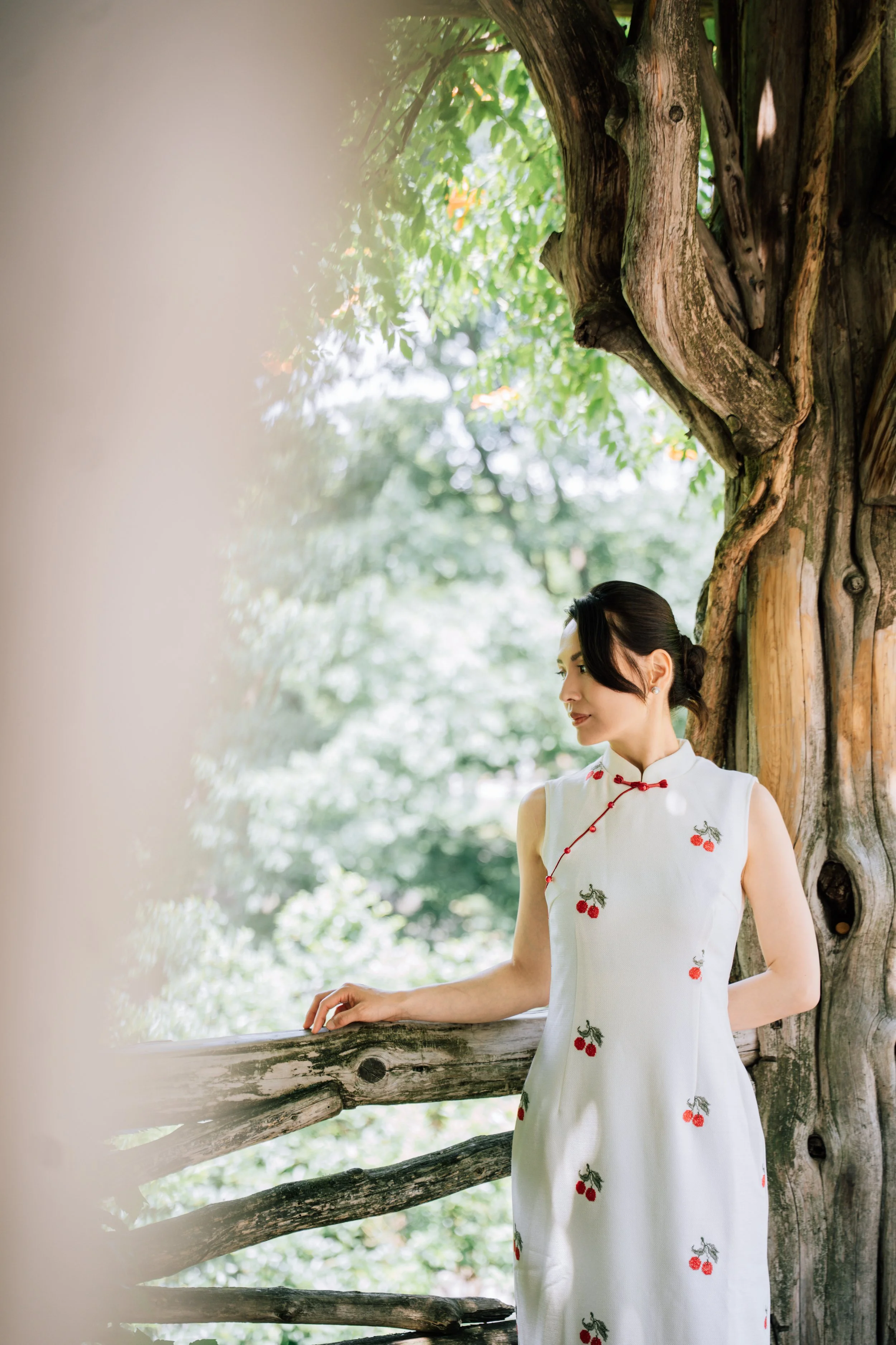 A woman in a white dress with cherry patterns standing outdoors by a wooden railing, with green trees and foliage in the background.